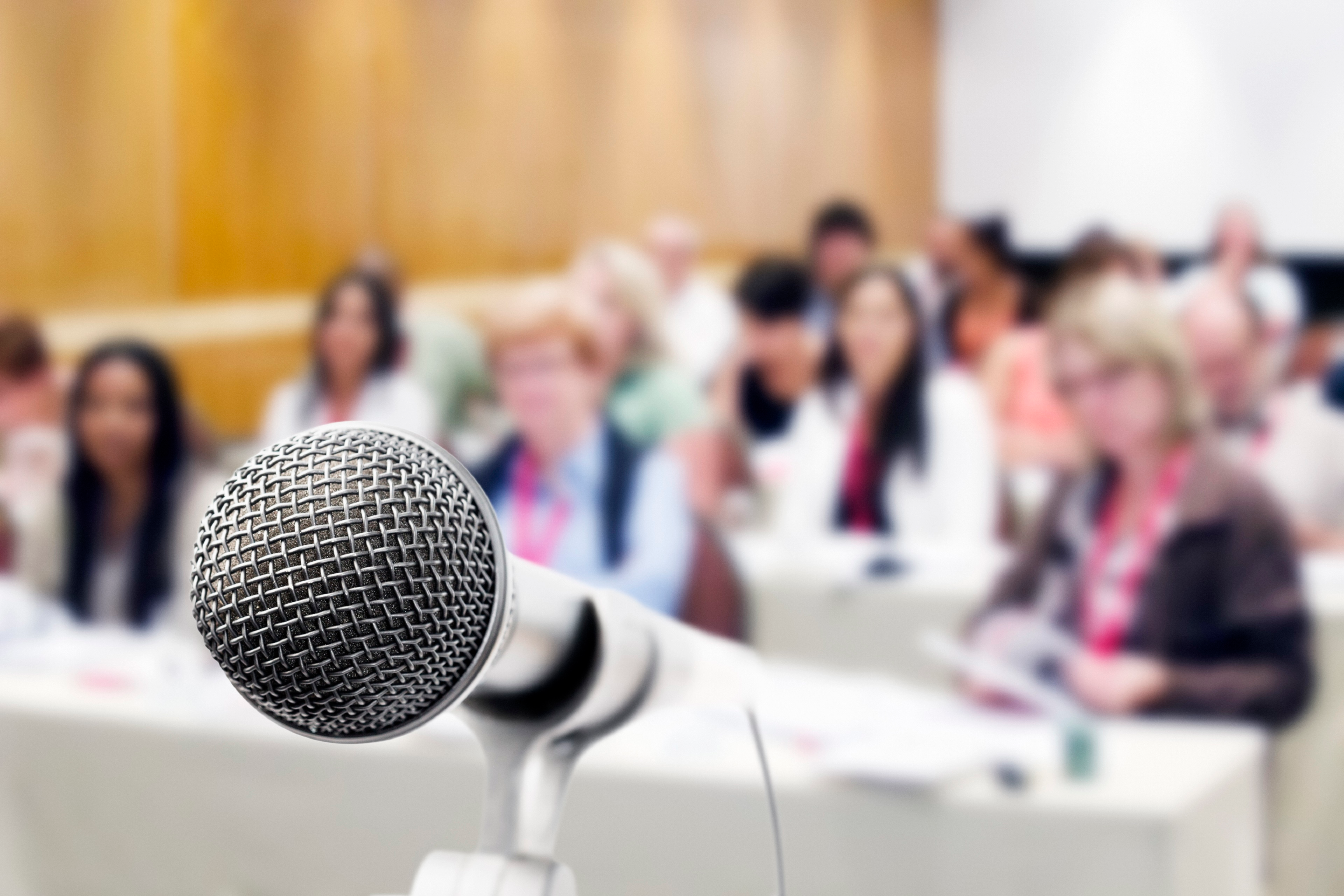 A vocal microphone stands ready against a background of an out-of-focus and unrecognizable audience at a meeting or seminar.