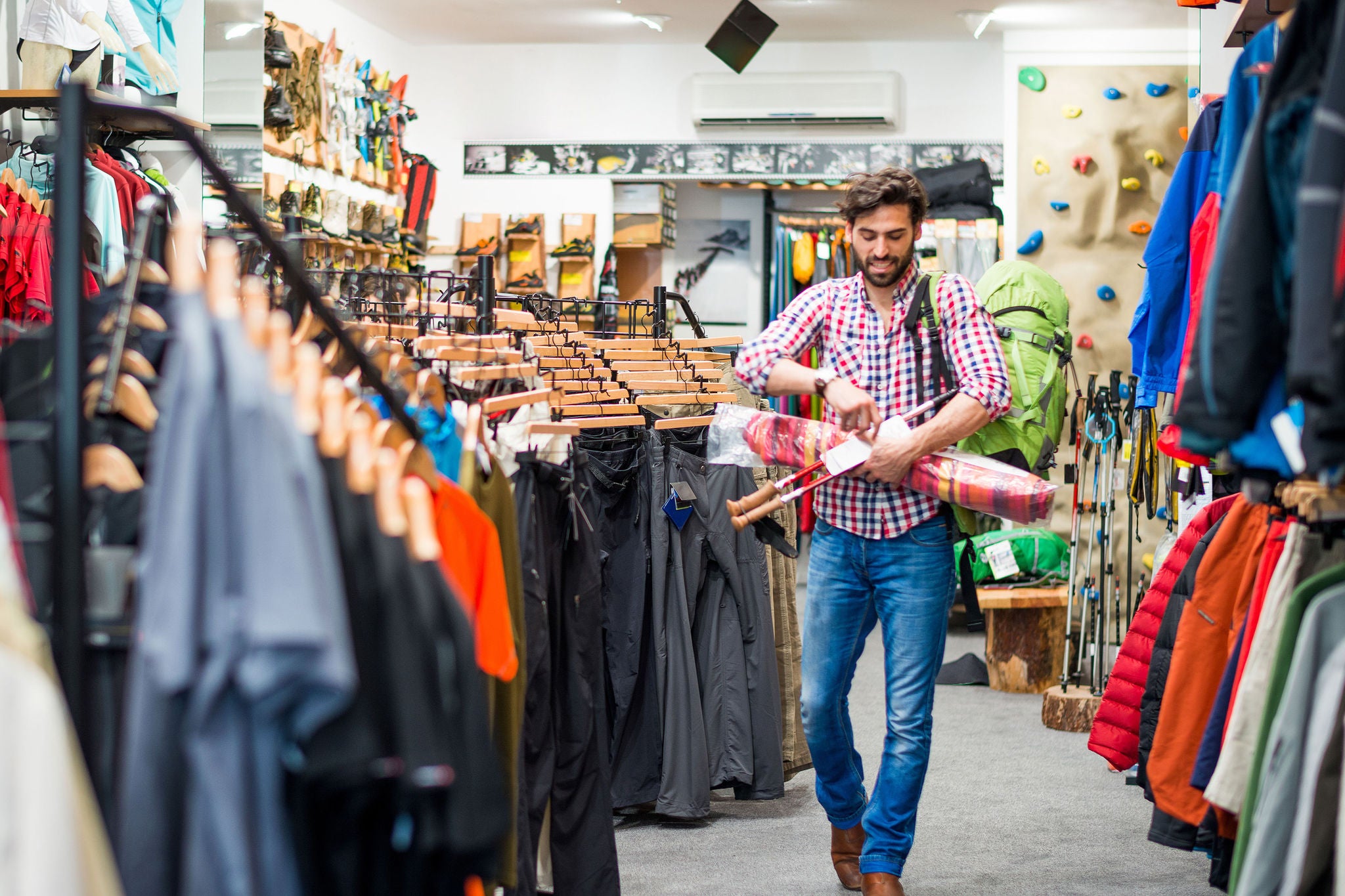 Bearded man in sports and outdoor equipment store looking to buy hiking equipment. He is holding green backpack, exercise mat, drinking bottle and hiking poles.