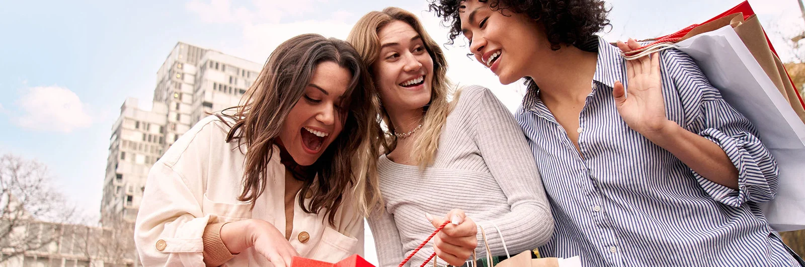 Three enthusiastic European friends on city street hold paper sack. Women coming from shopping in the sales look at inside the recyclable bags surprised. Smiling amazed people addicted to purchases.