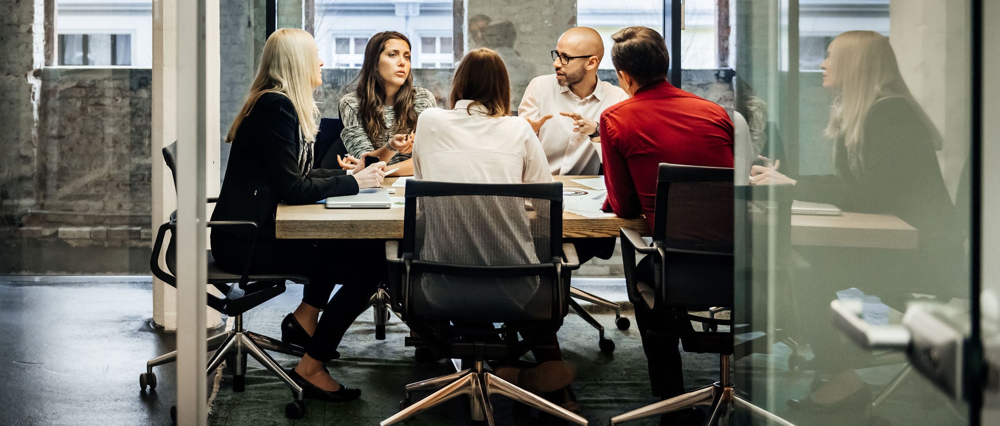 A group of people is sitting at a table during a business meeting in a bright, modern office. The team is talking business while pie charts can be seen hanging on the wall. Big bright windows are seen in the background.