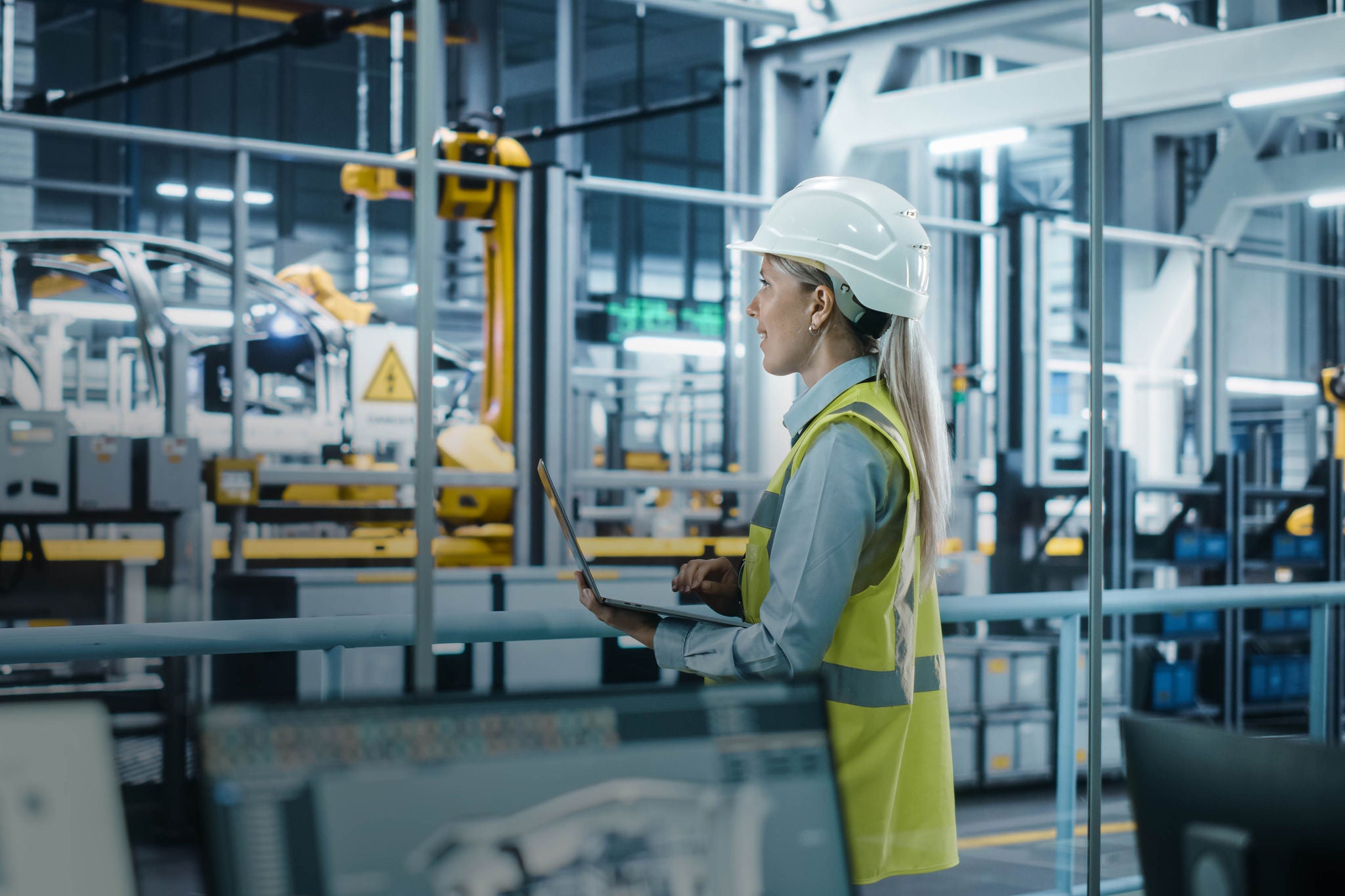 Car Factory: Female Automotive Engineer Wearing Hard Hat, Standing, Using Laptop. Monitoring, Control, Equipment Production. Automated Robot Arm Assembly Line Manufacturing Electric Vehicles.