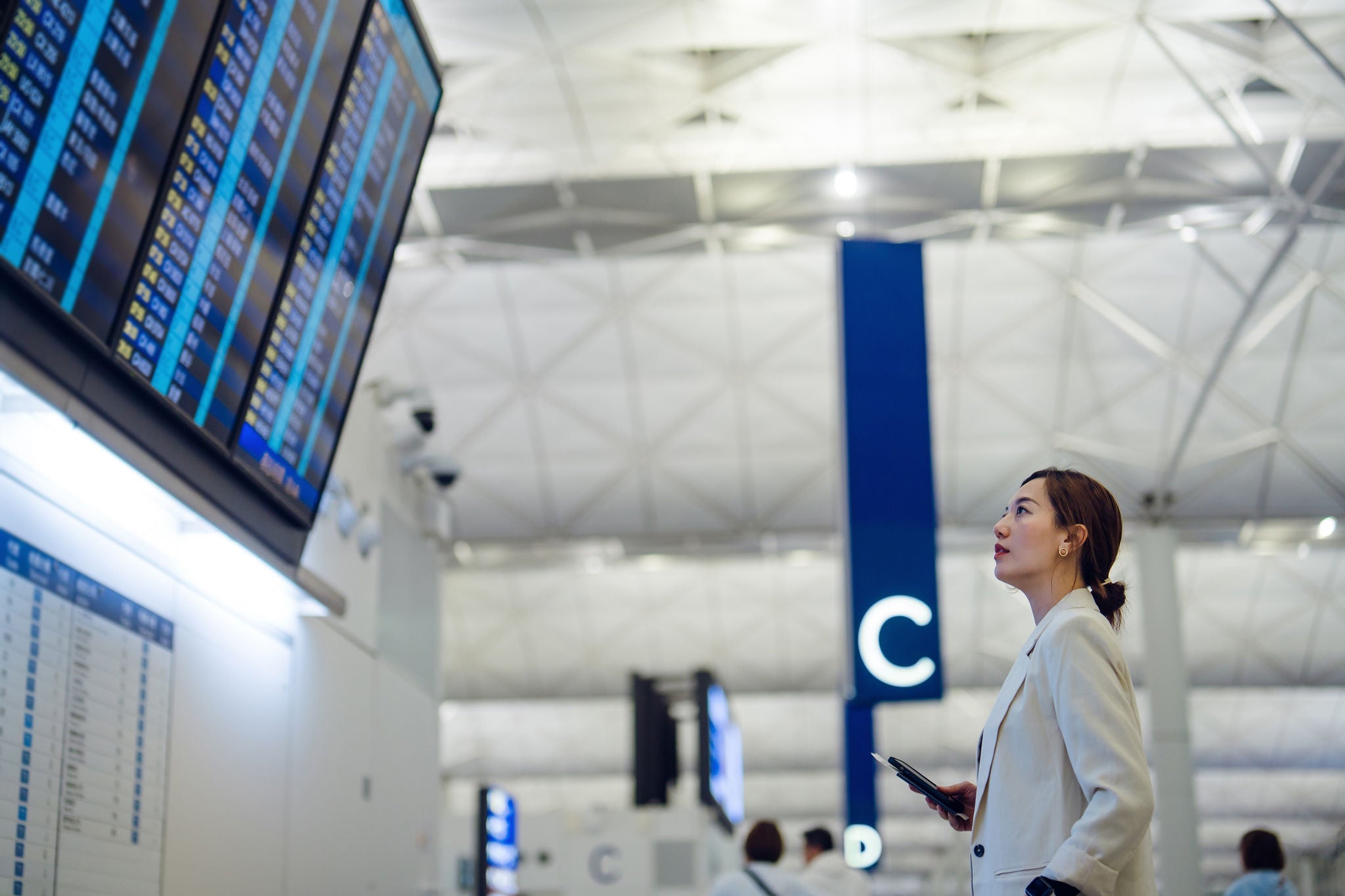 Young Asian businesswoman with passport and boarding pass, looking up and checking her flight schedule from the Arrival Departure Board at airport terminal. Business trip. Business travel. Travel and vacation concept