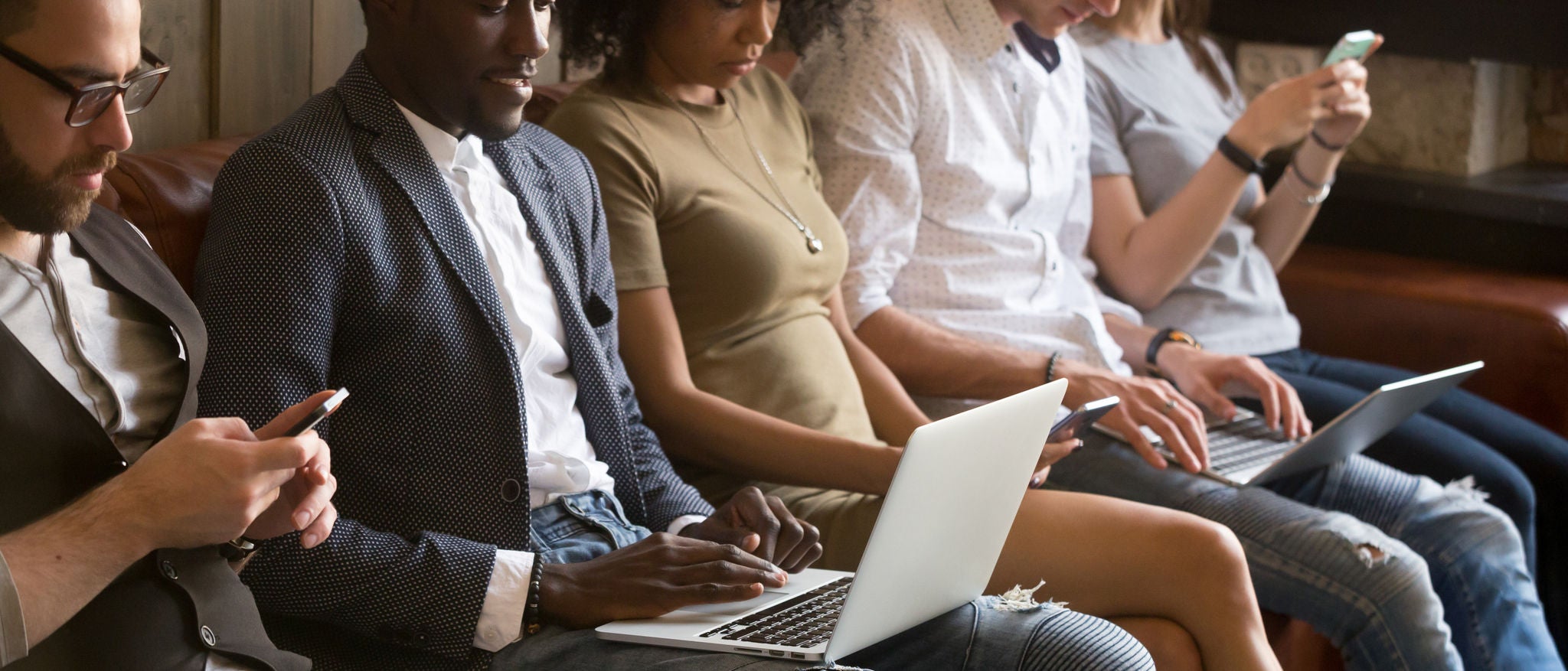 Close up of diverse young people being addicted to gadgets, sitting in queue using phones and laptops, multiracial millennials busy chatting or checking email on smartphones, obsessed with technology