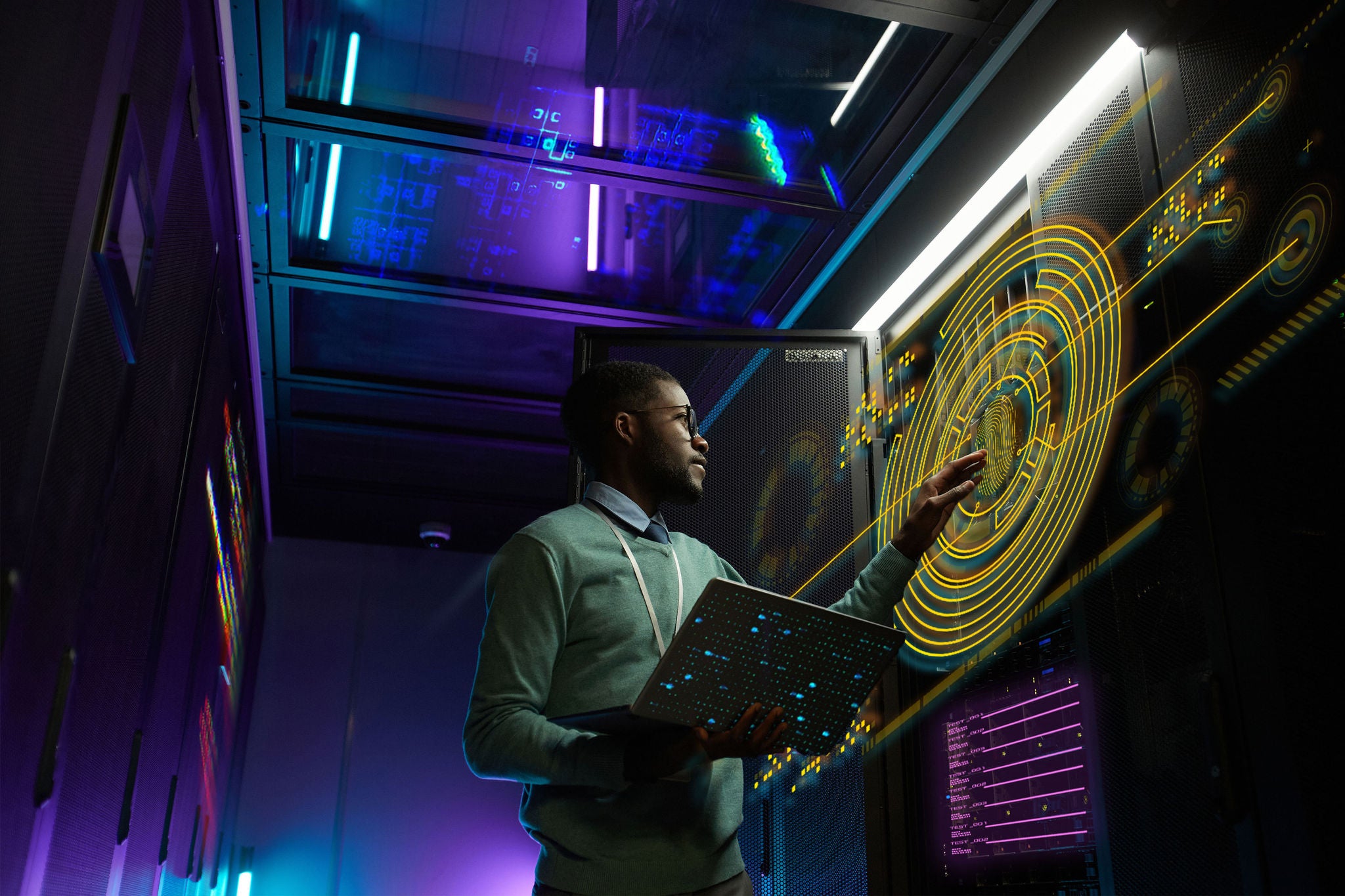 Low angle portrait of young African American data engineer working with supercomputer in server room lit by blue light and holding laptop, copy space