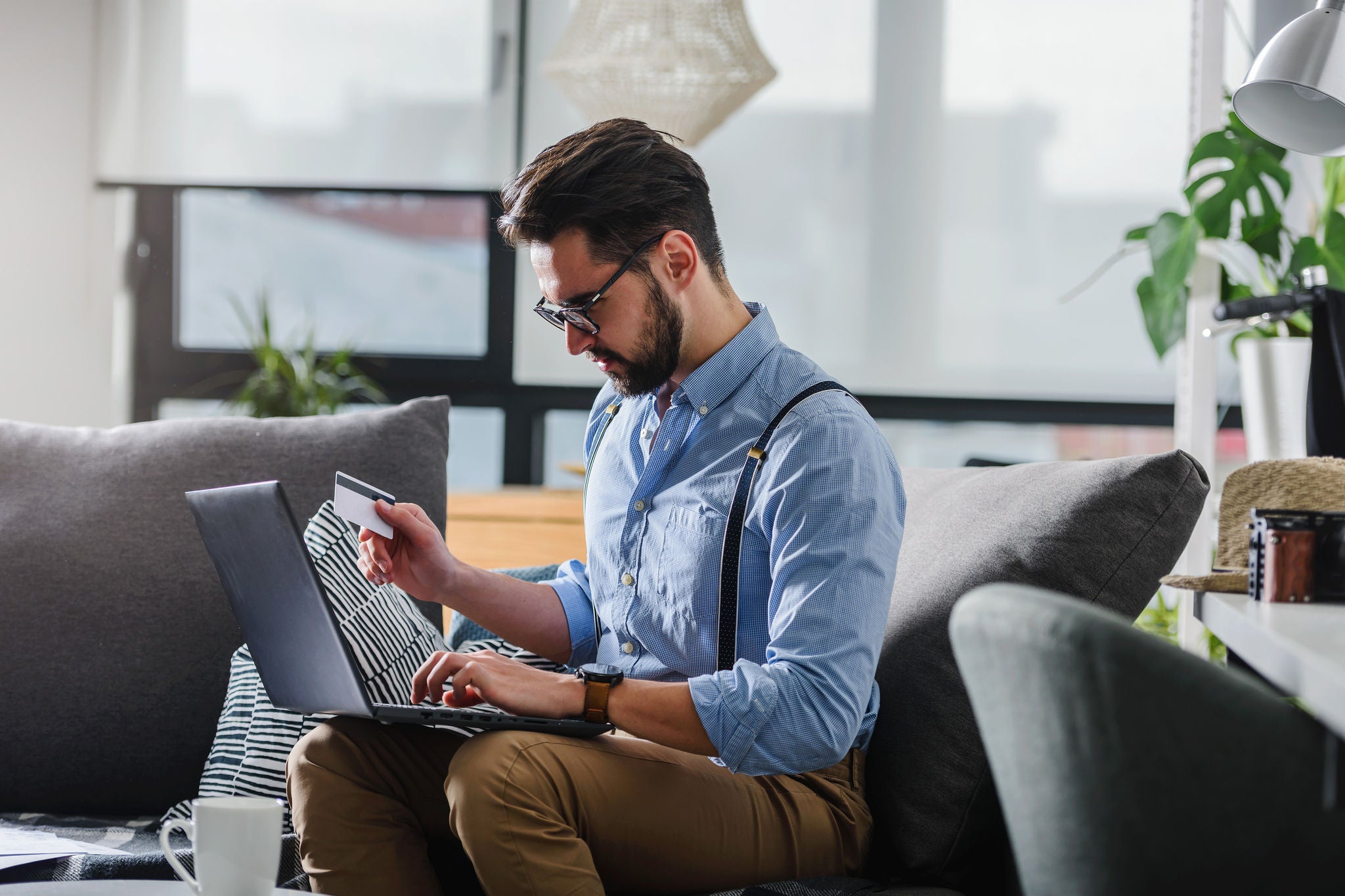 A man wearing glasses and a light blue shirt sits on a couch in a modern living room, focused on his laptop. 