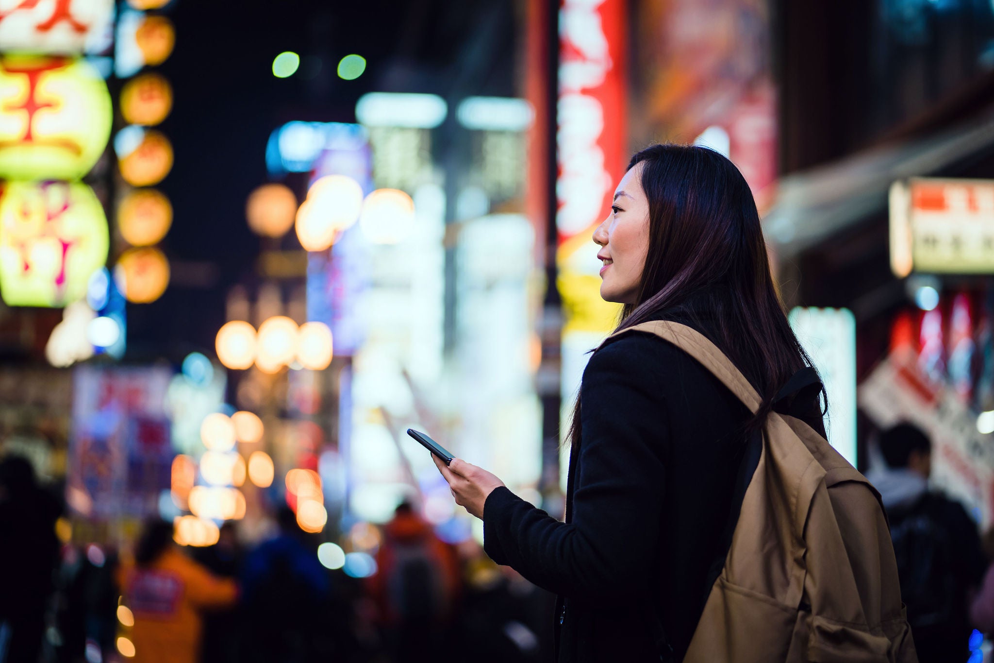 Young Asian female traveller with backpack using smartphone while exploring and strolling in busy local city street at night in Osaka, Japan