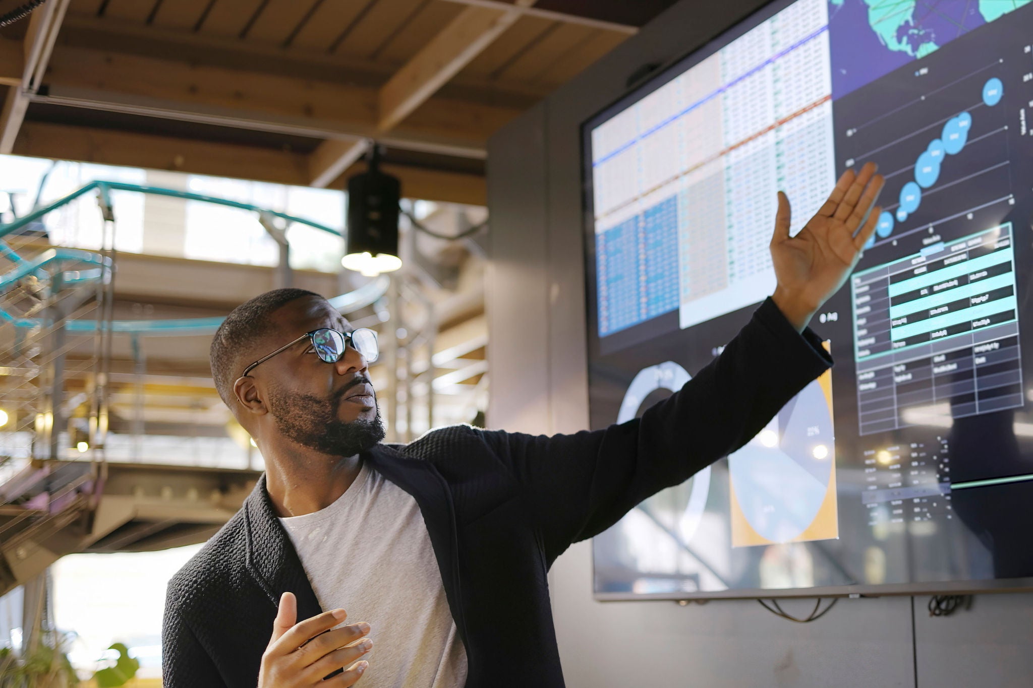 Stock image of a Black man conducting a seminar / lecture with the aid of a large screen. The screen is displaying graphs & data associated with movie clips of the earth.