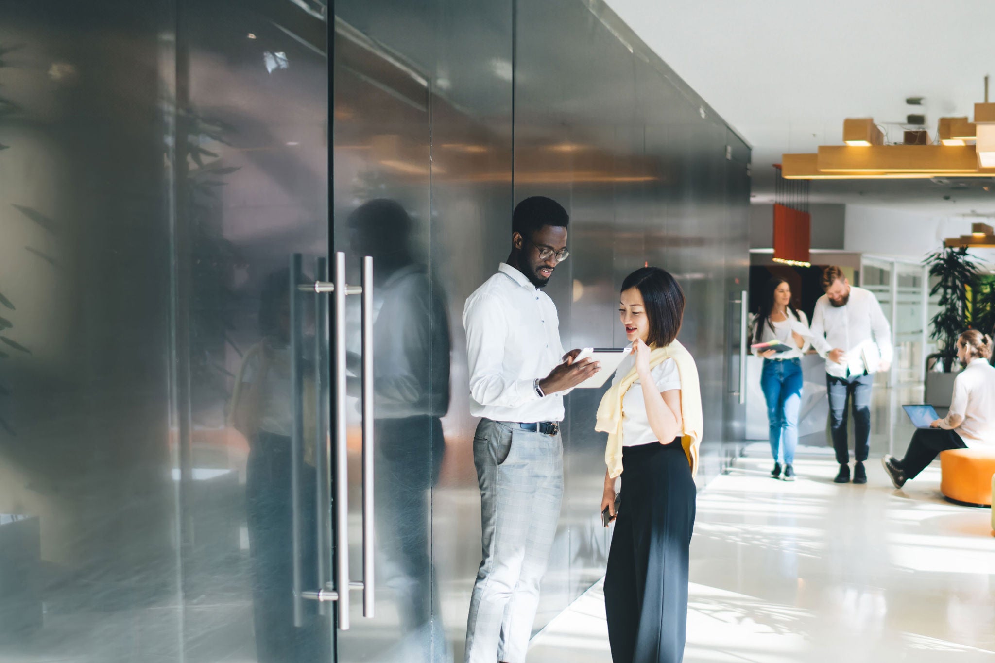 Multiethnic company staff in casual clothes walking and standing in big sunlit corridor of modern workspace discussing business and working with gadgets