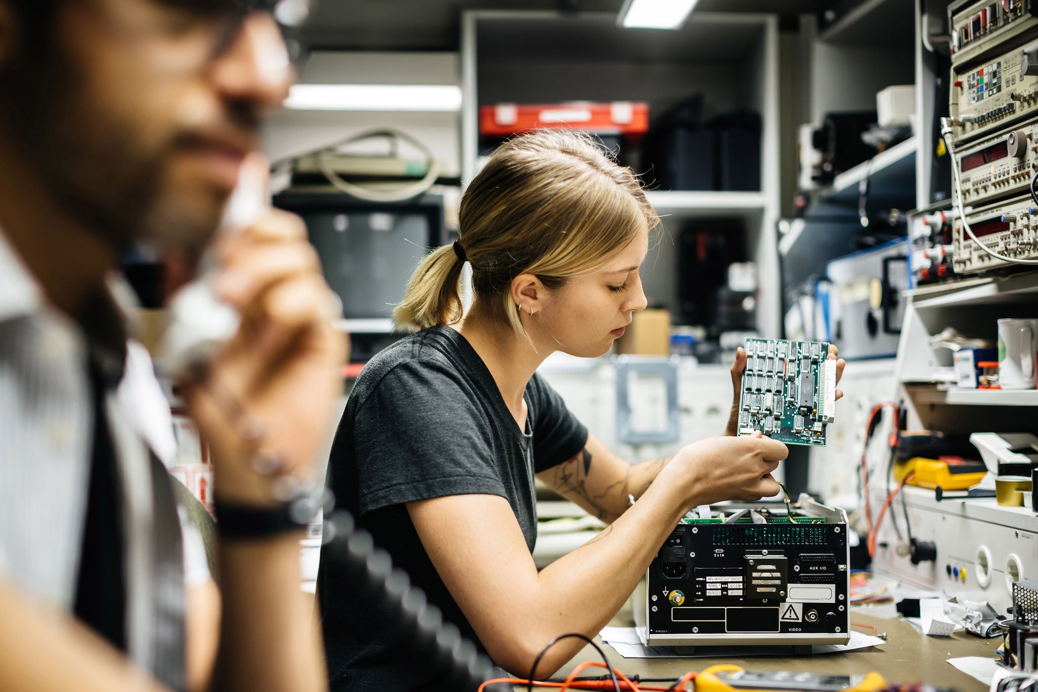 Female Technician working on a conductor board in workshop. Her male colleague calling in foreground
