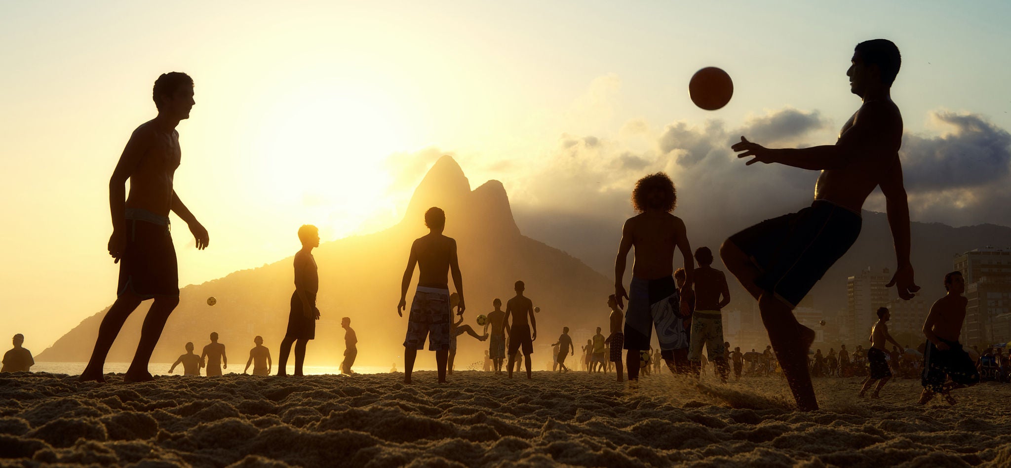 Silhuetter av fotballspillende personer på en strand, omgitt av varme solstråler i en idyllisk solnedgangsscenario.