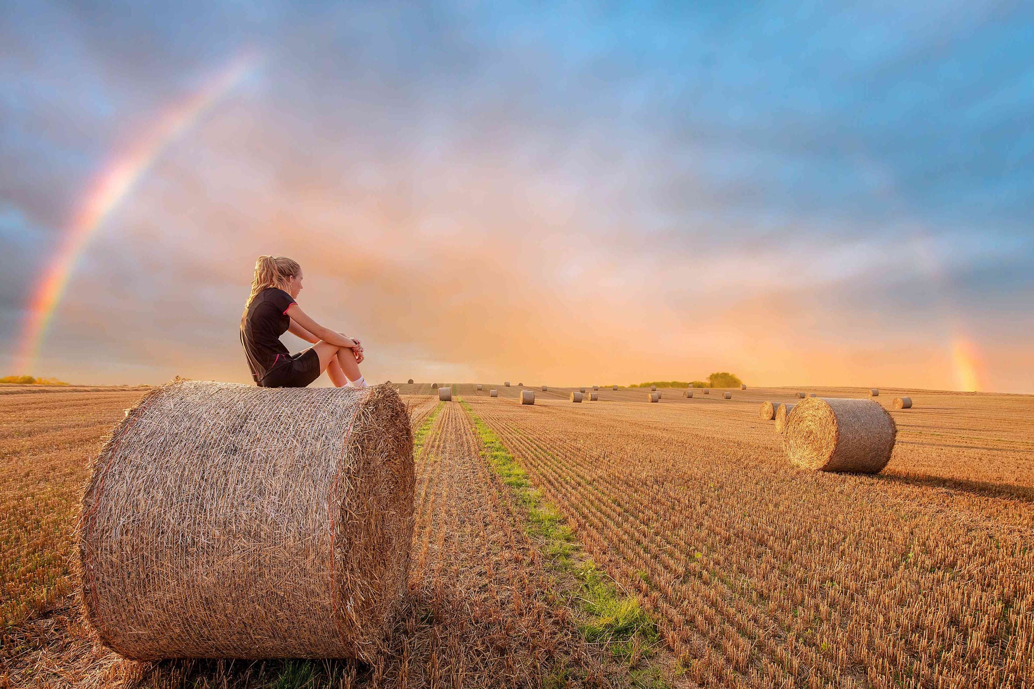 Woman looking sat on hay bales as a rainbow appears after a rain shower