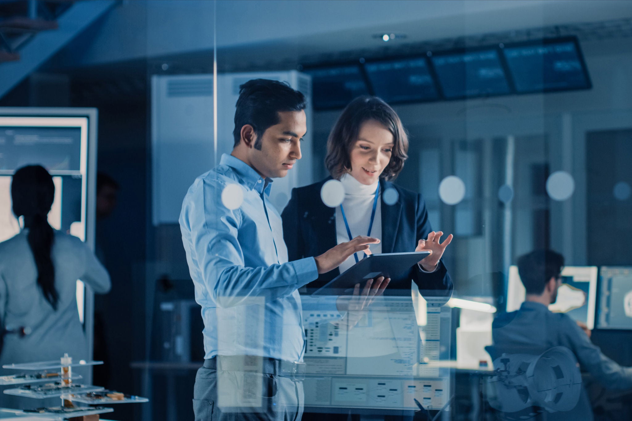 In Technology Research Facility: Female Project Manager Talks With Chief Engineer, they Consult Tablet Computer. Team of Industrial Engineers, Developers Work on Engine Design Using Computers