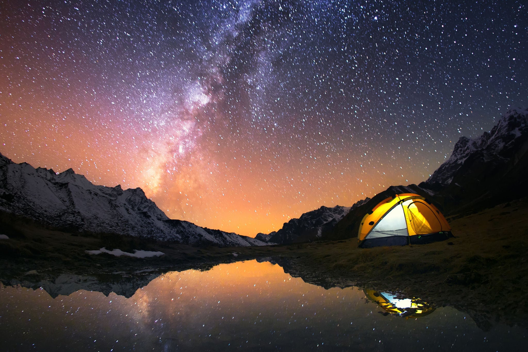 漫天星空與群山環繞下湖畔一個亮著燈的露營帳篷。