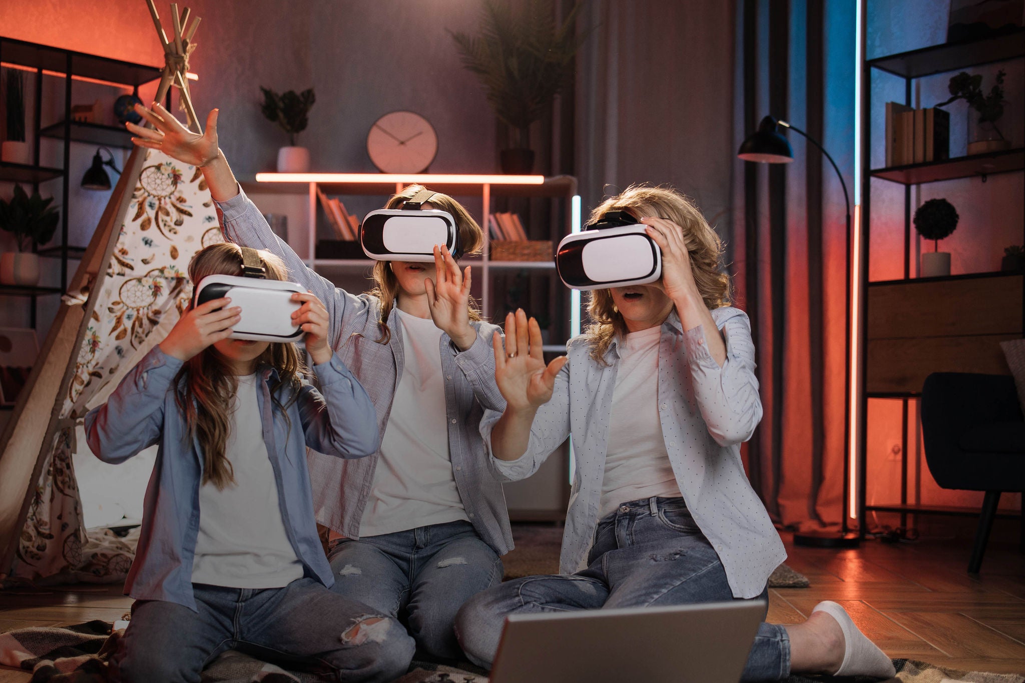 Happy mother and cute daughters sitting near toy wigwam at home and using modern VR headset during evening time. Caucasian family using modern technologies for playing at home.