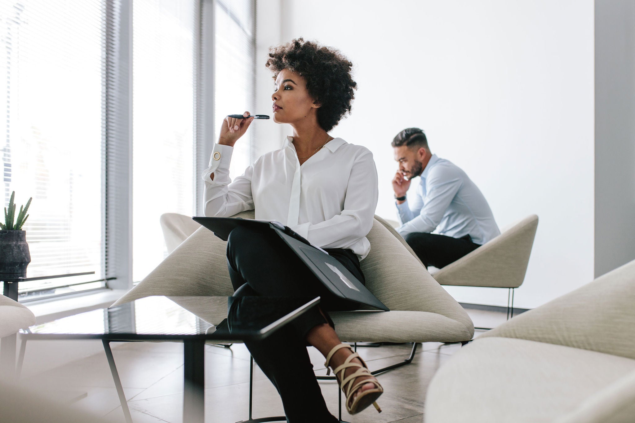 Young woman sitting in office hallway with a file and looking away thinking. Business woman thinking over a new business proposal with a coworker talking on mobile phone in background.