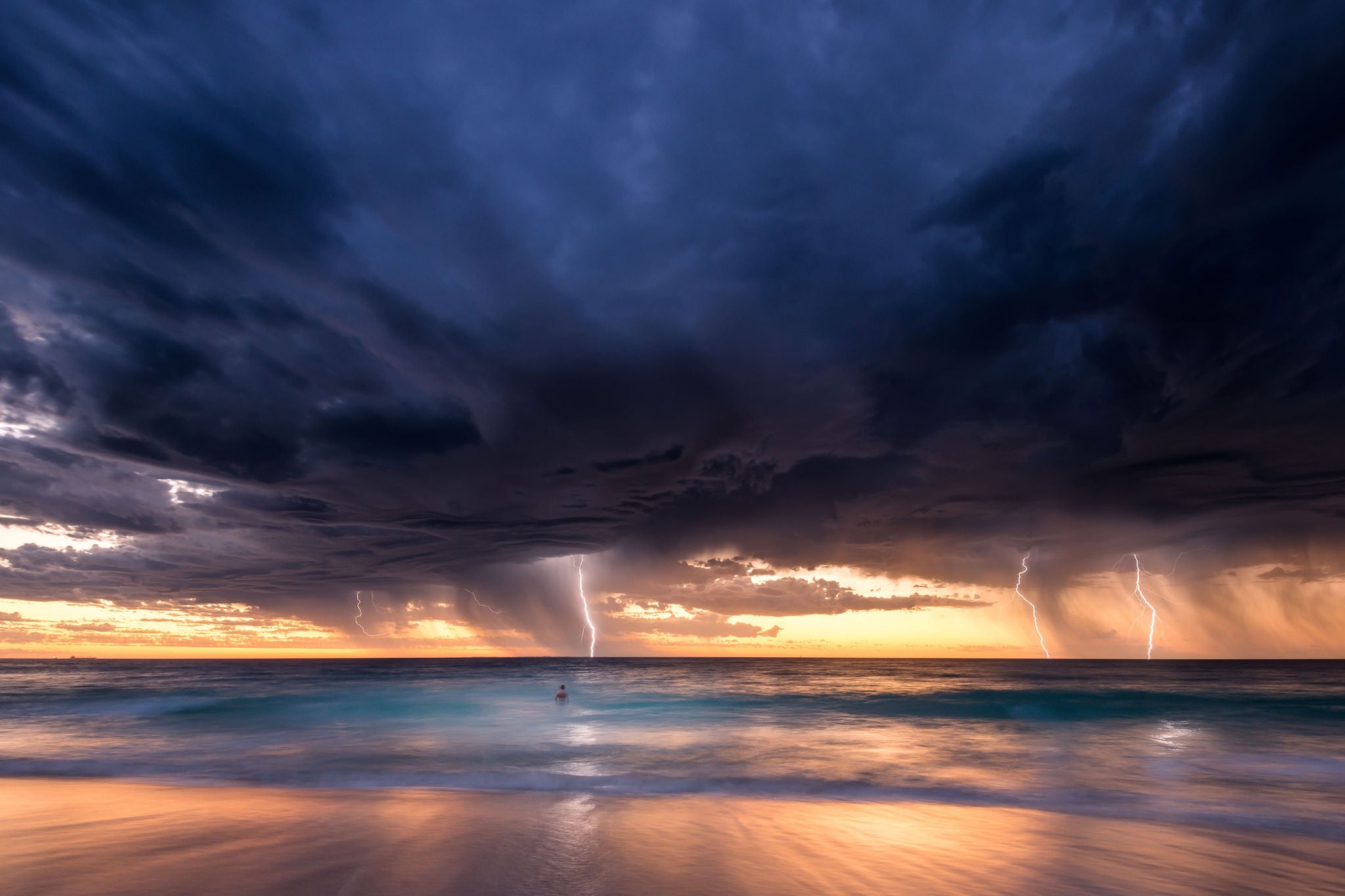 A dramatic coastal scene featuring a dark stormy sky with thick clouds looming over a beach at sunset