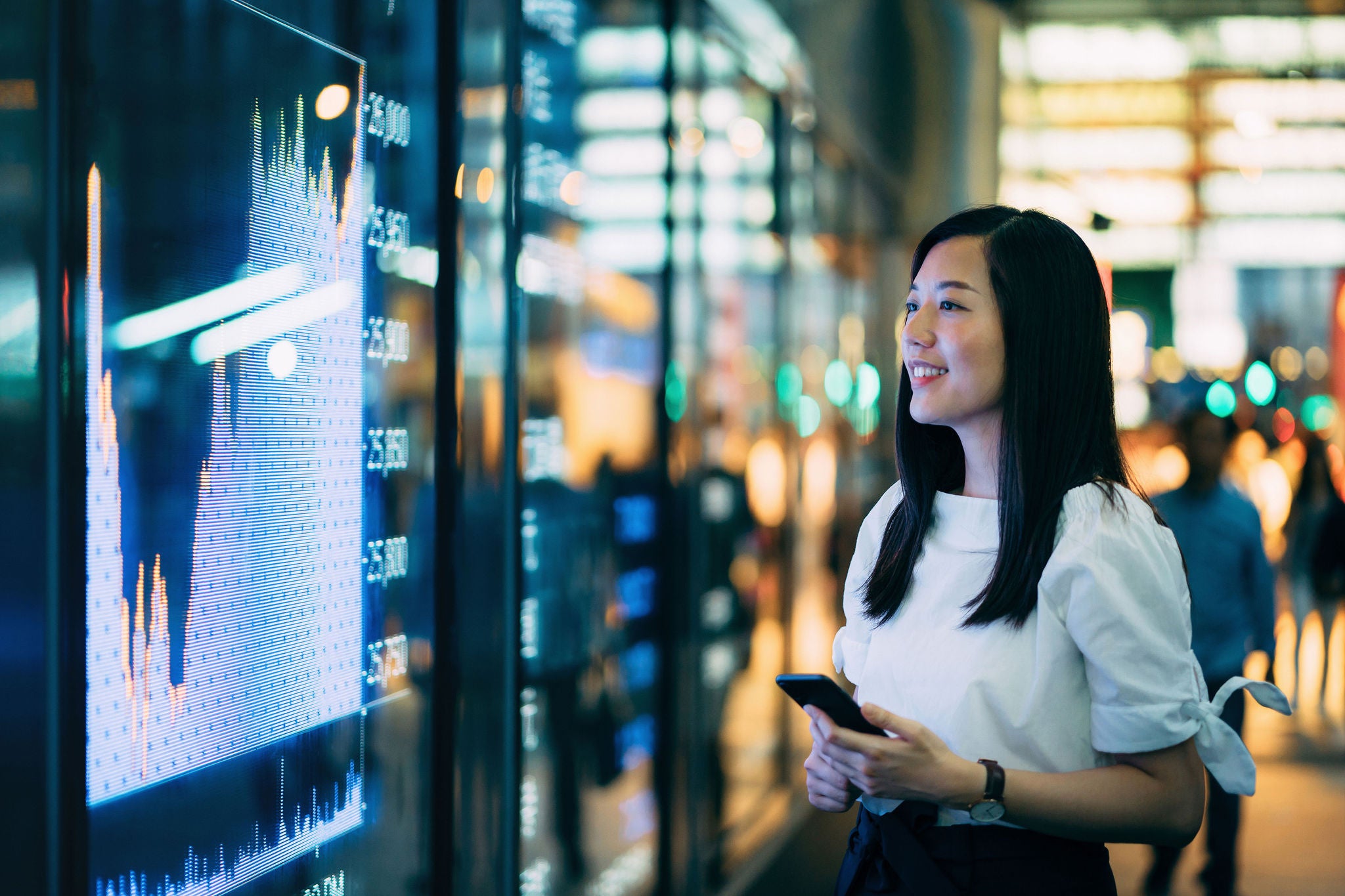 Asian businesswoman on smartphone by the stock exchange market display screen