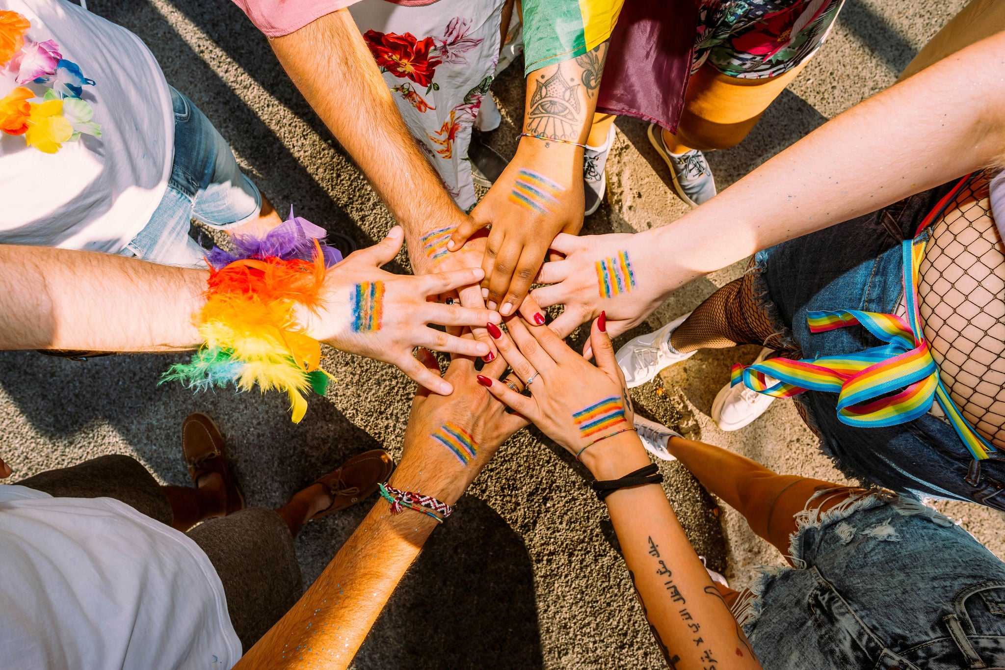 Eine Gruppe von diversen, multikulturellen AktivistInnen mit Regenbogenaccessoires stehen im Kreis und kreuzen ihre Haende