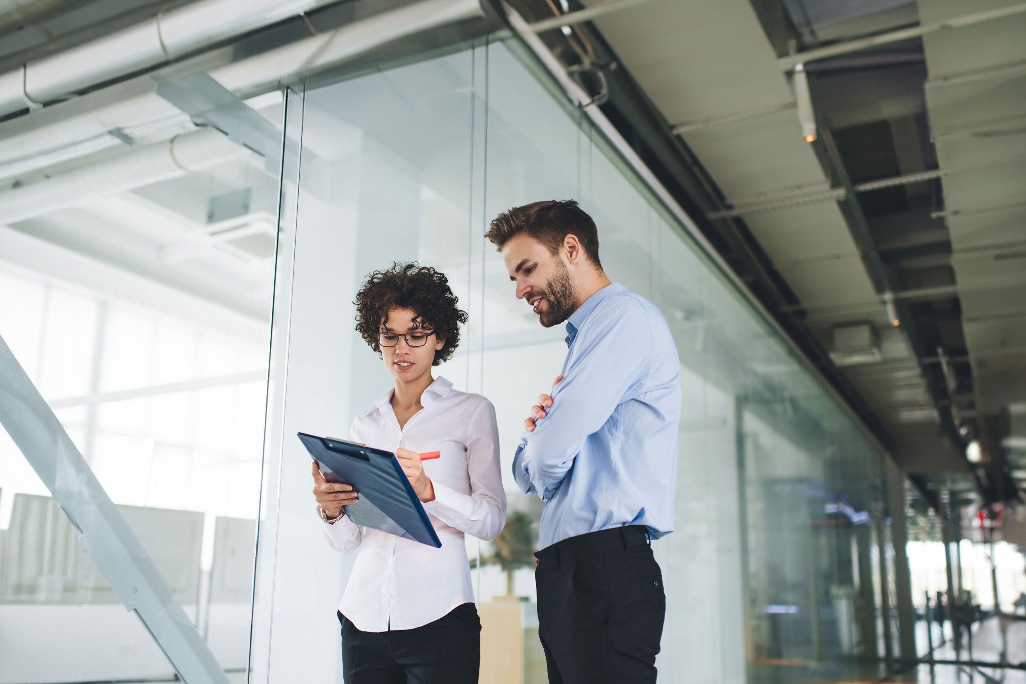 Businessman with crossed arms looking what his female colleague writing on clipboard. Business cooperation and teamwork. Young caucasian millennial colleagues in office. Modern successful people