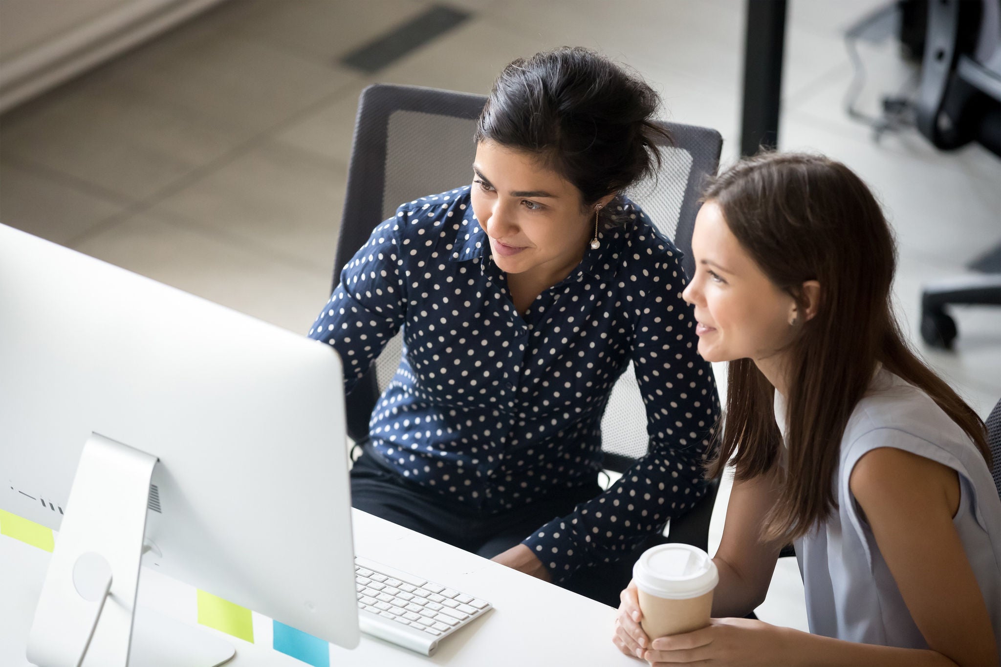 Two women working together in a modern office setting, closely looking at a computer screen.