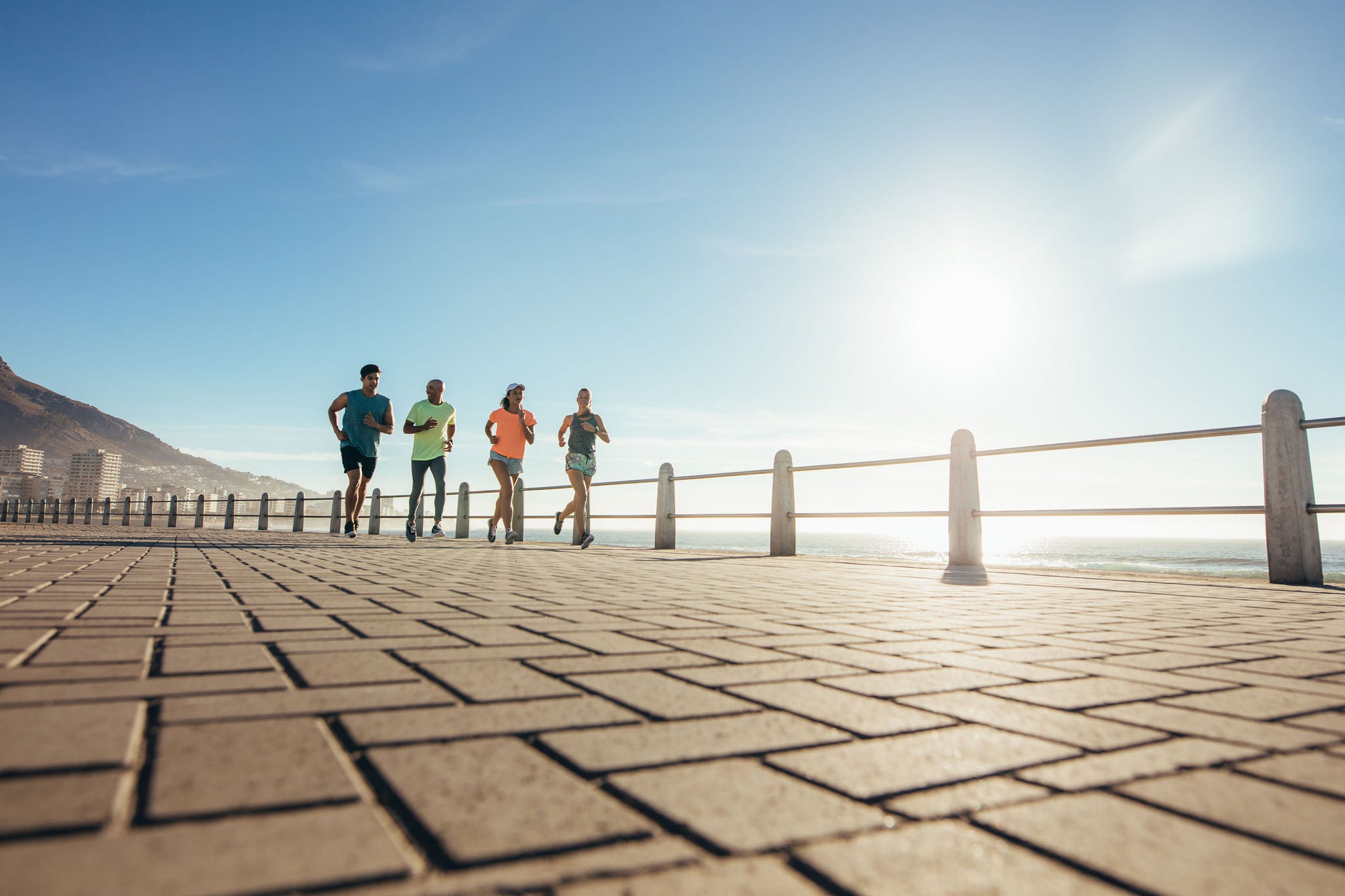 Low angle shot of four young people running on ocean water front. Running on seaside road on a sunny day.