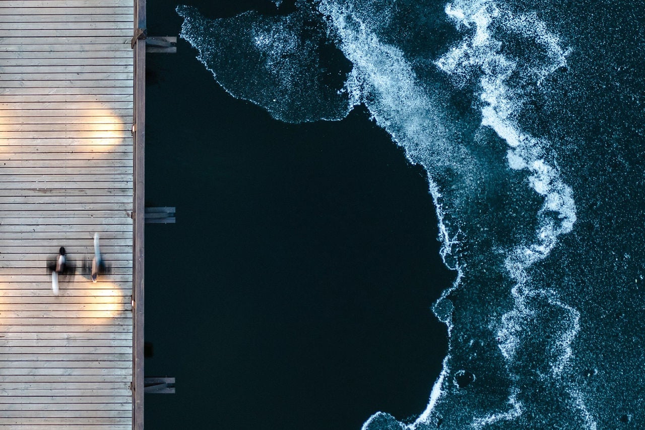 Top-down view of a wooden pier with blurred figures walking, next to a body of water with waves and ice formations along the shoreline.