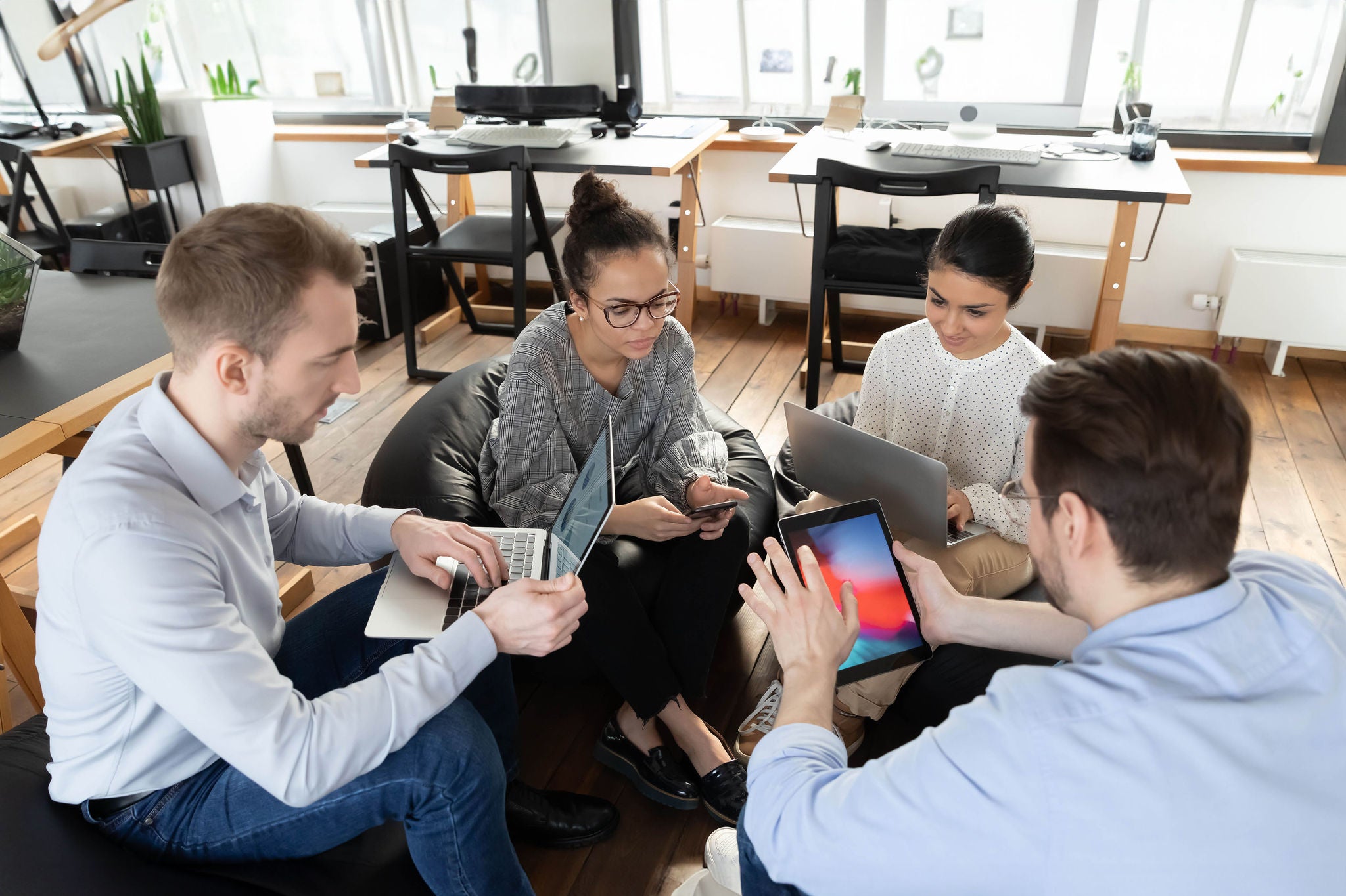 Focused multiracial colleagues sit in circle in office work on modern gadgets during casual meeting.