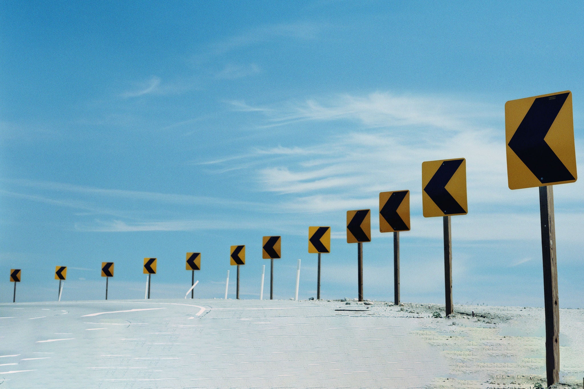 A row of yellow traffic signs with black left-pointing arrows positioned along a curved road under a clear blue sky.