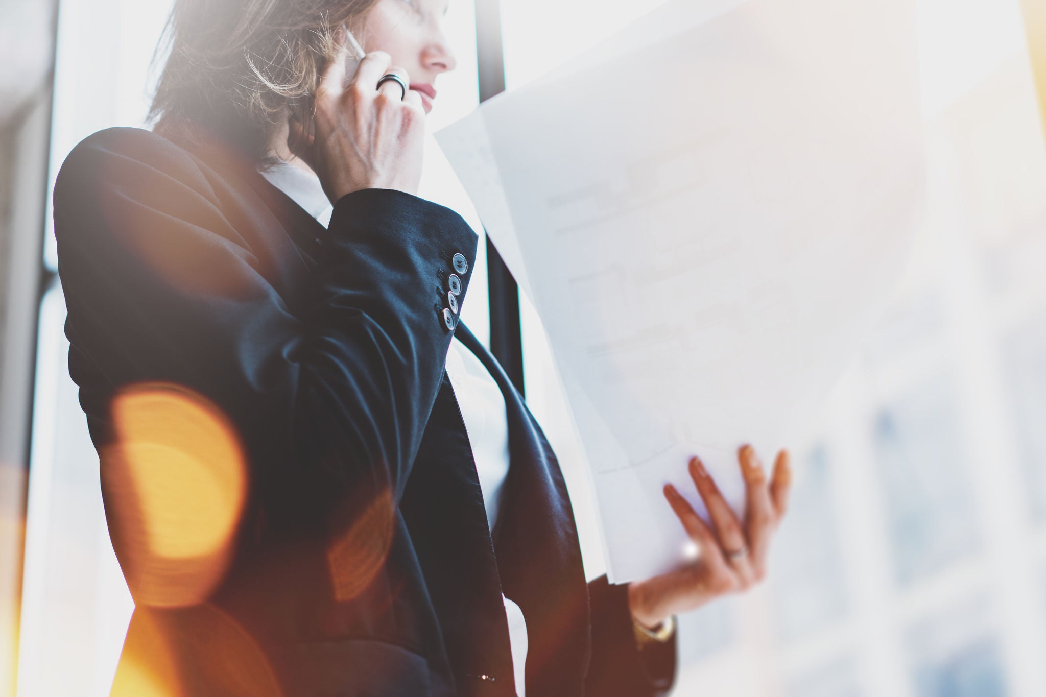 Photo business woman wearing white shirt, talking smartphone and holding documents in hands. Open space loft office. Panoramic windows background. Wide mockup