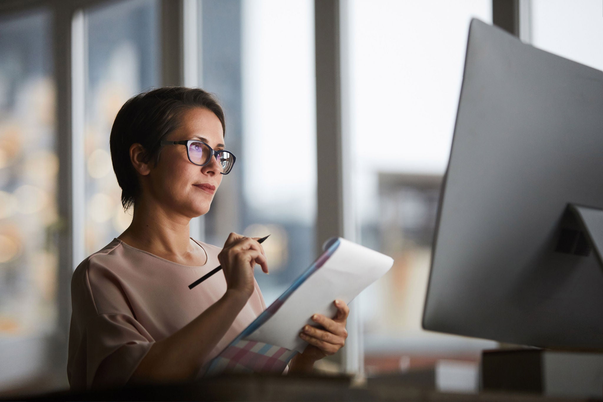 Confident businesswoman with document looking at computer screen and making working notes in office