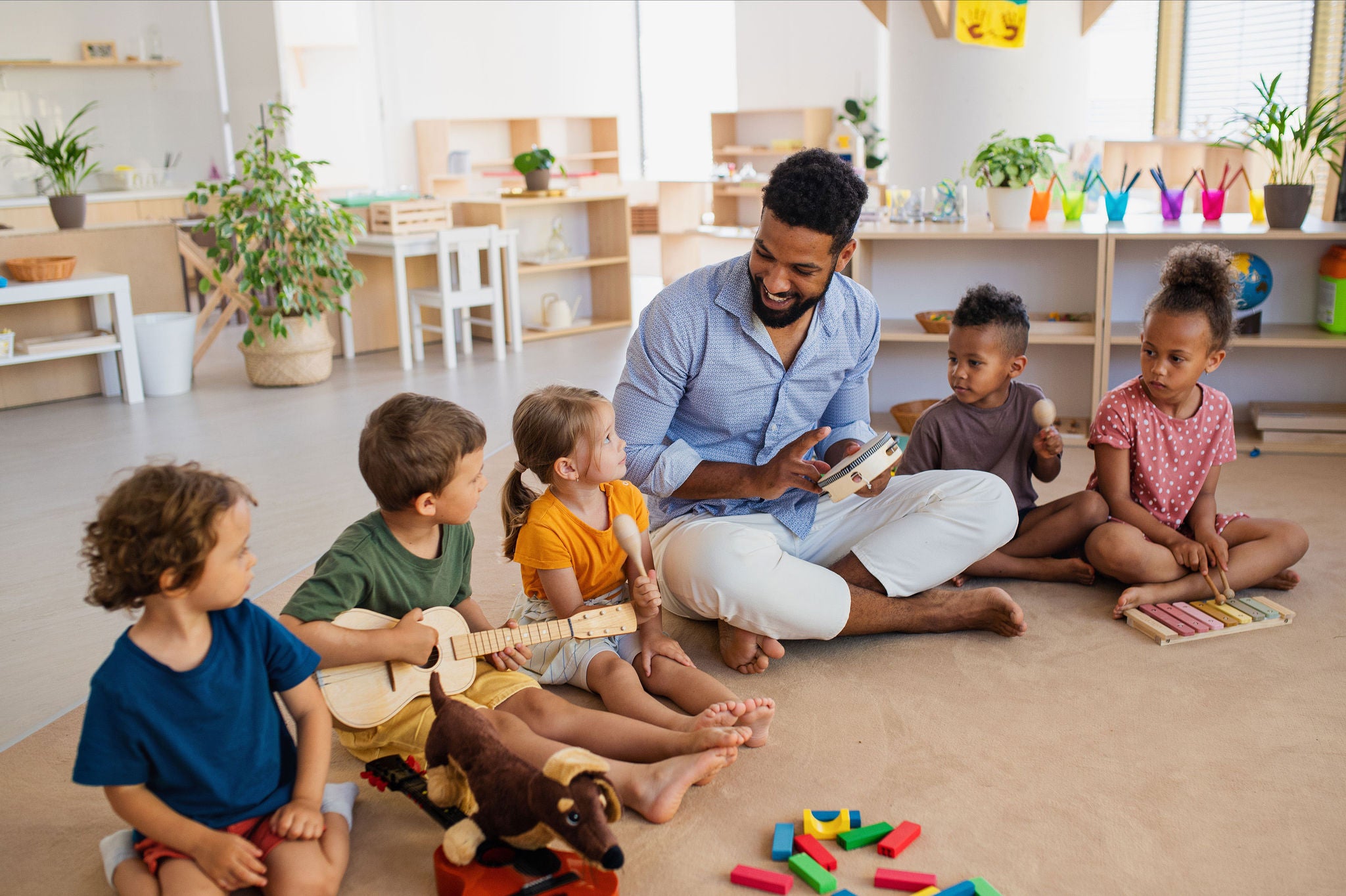 A group of small nursery school children with man teacher sitting on floor indoors in classroom, playing musical instruments.