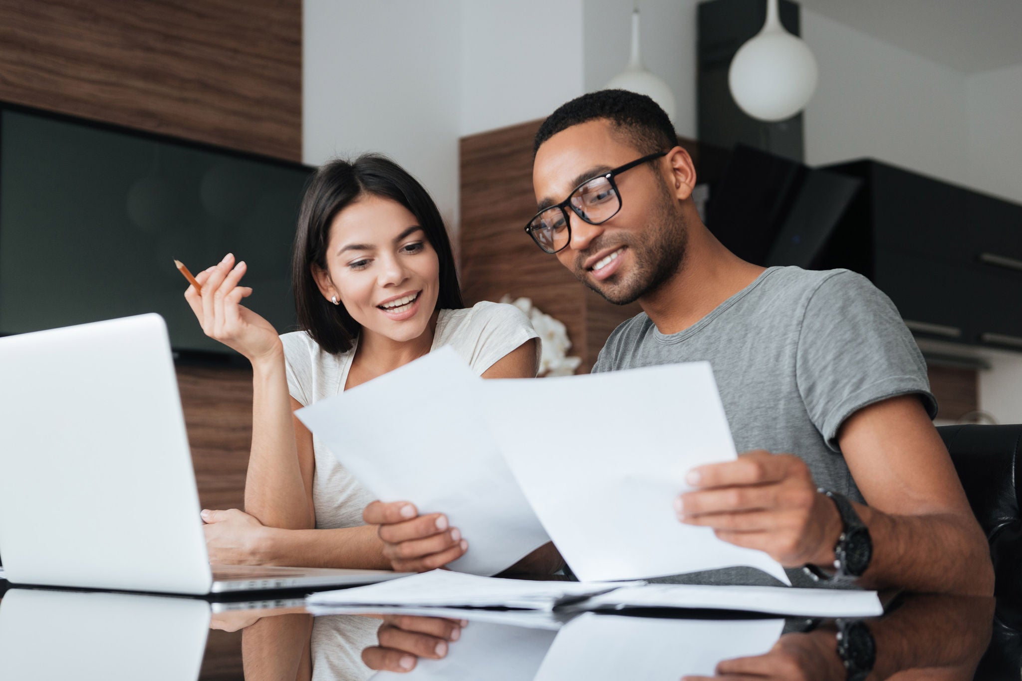 Photo of cheerful loving young couple using laptop and analyzing their finances with documents. Look at papers.