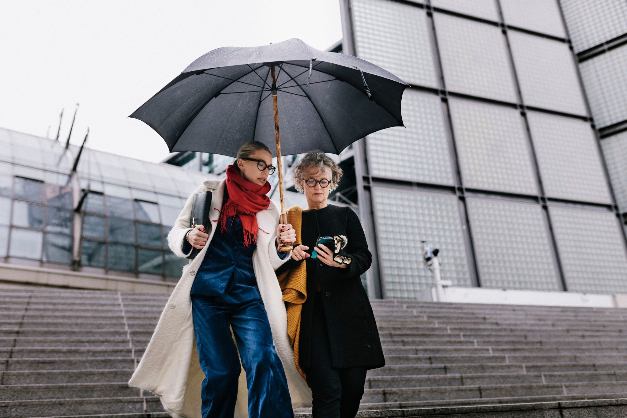 A female CEO walking outdoors in the rain with her assistant, who is holding an umbrella and protecting her while she uses a smartphone.