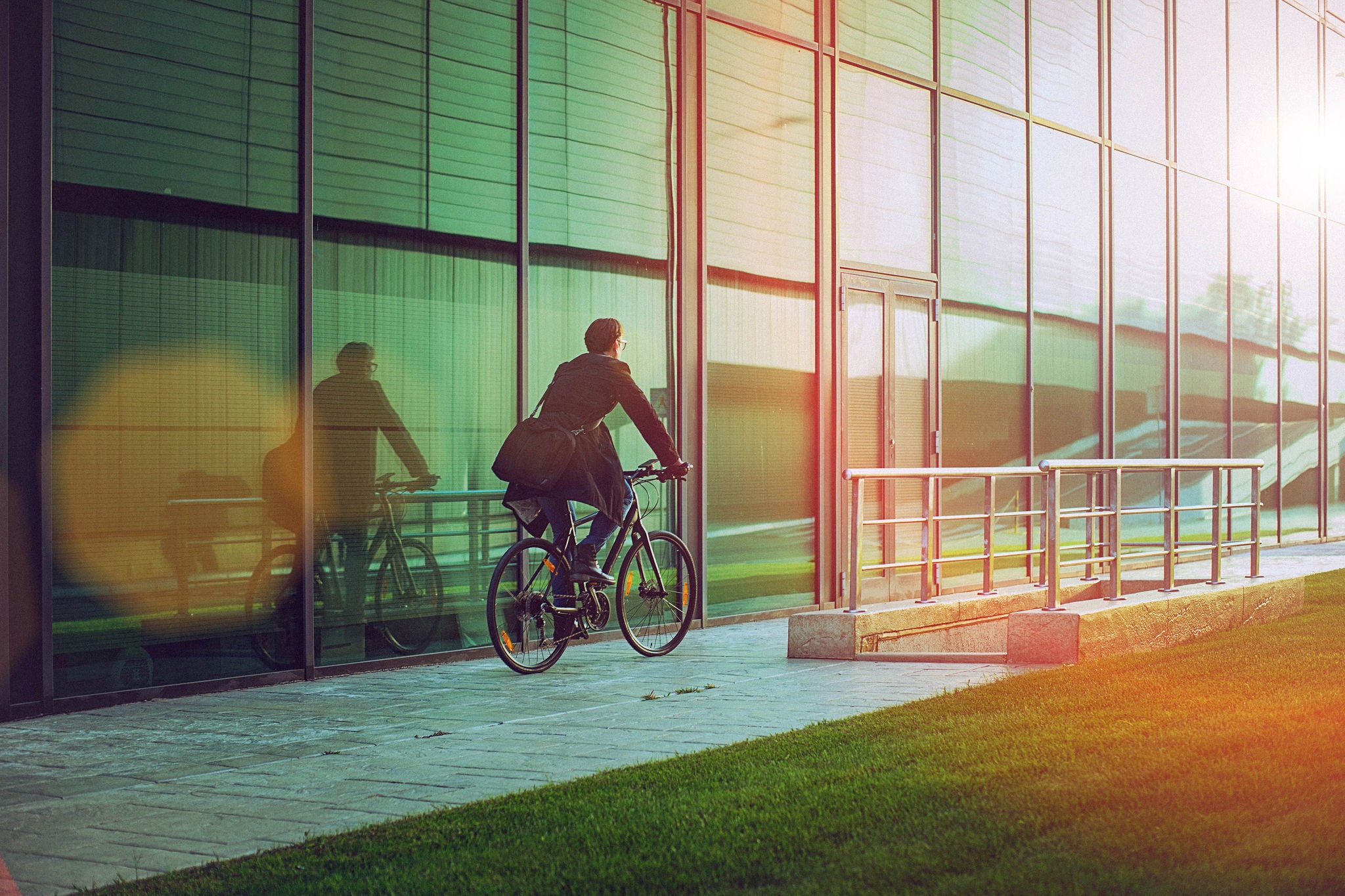 Rear view of a handsome man, on the way to work, riding bicycle beside the modern office building. The man is casually dressed and wears eyeglasses and carries black briefcase hung on shoulder. Blurred motion, copy space has been left.