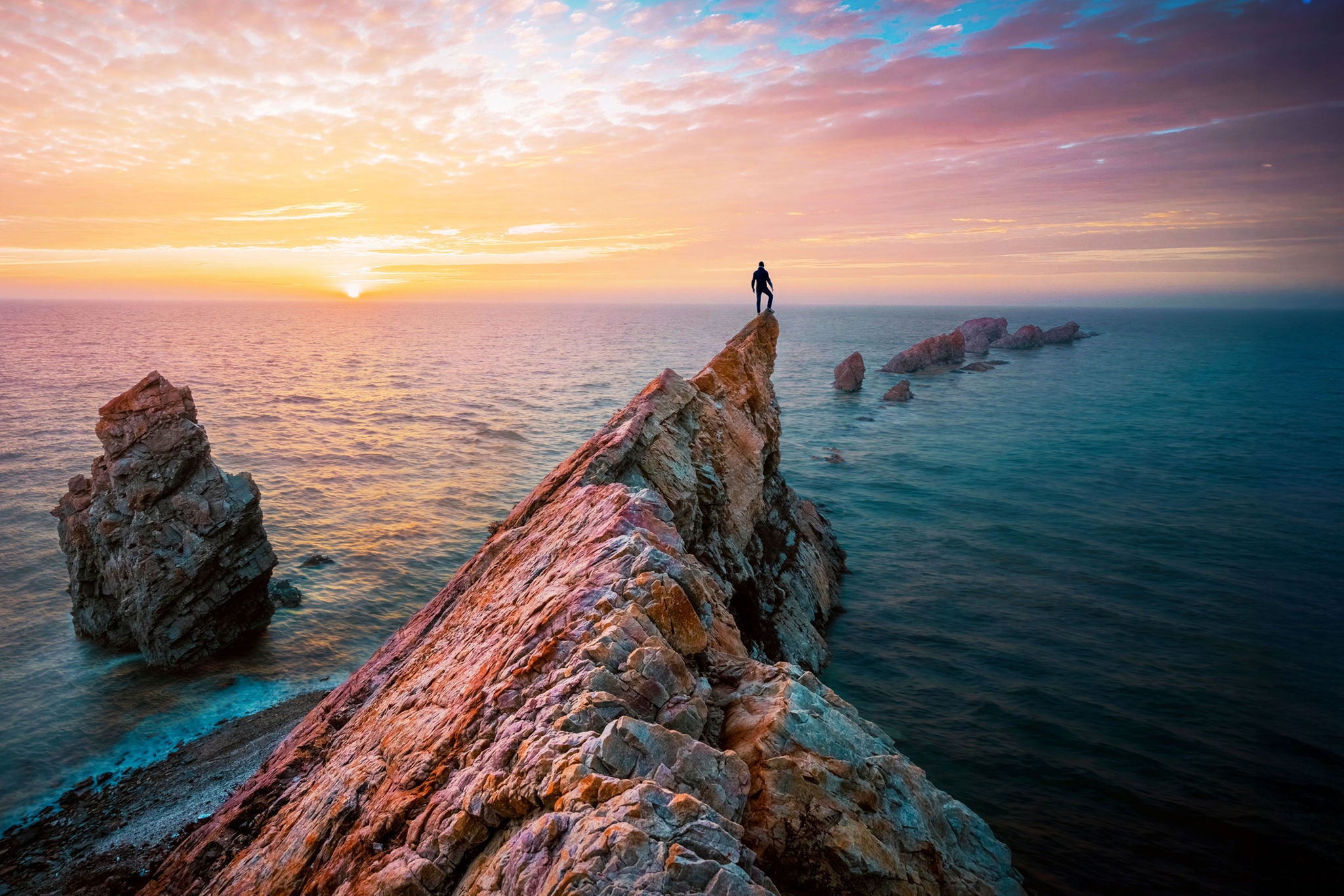 Man standing on rocky outcrop by sea watching sunset