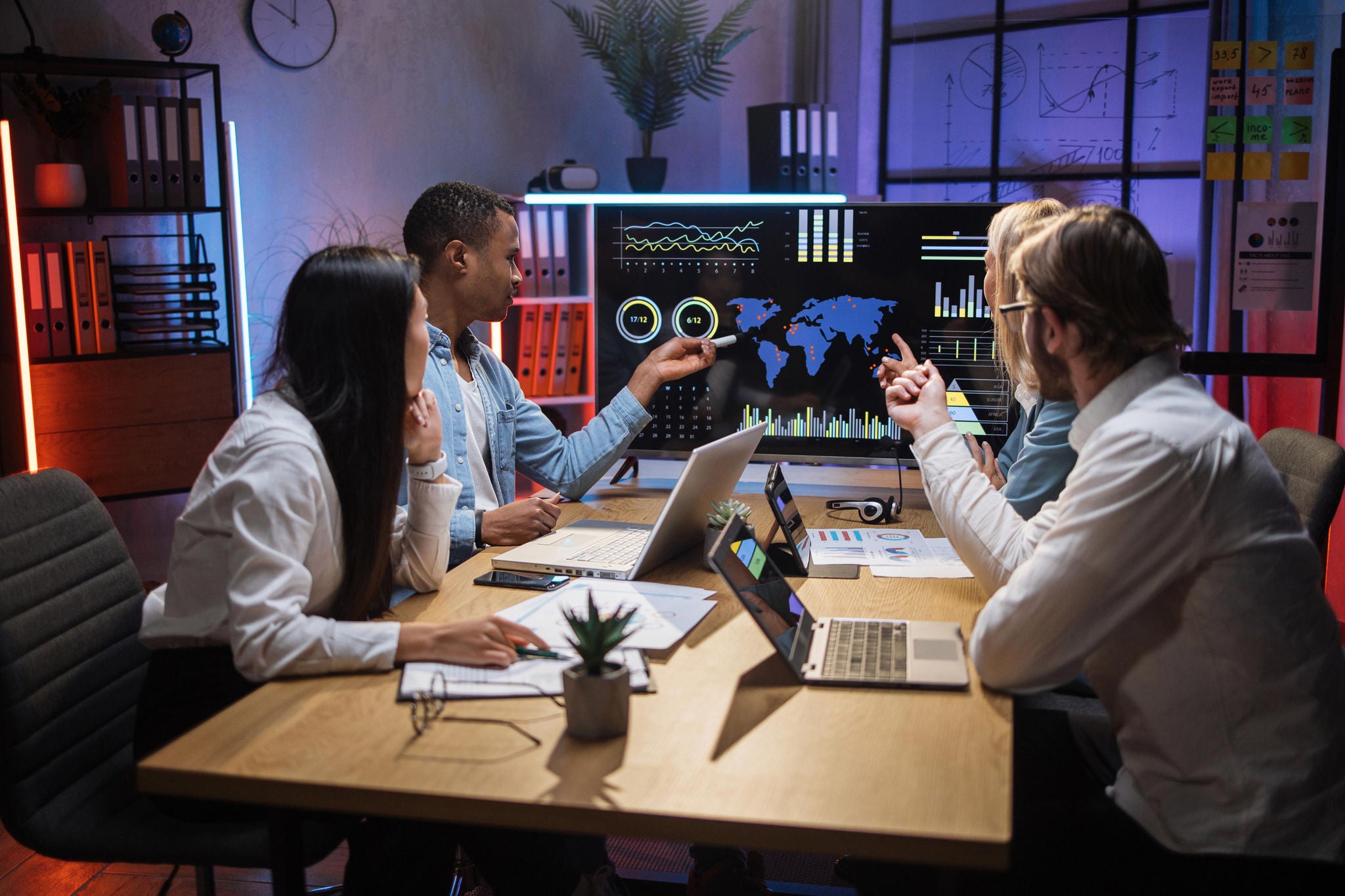 Multi ethnic team of IT workers spending evening time at office for making strategy of successful software project. Four people sitting at desk and looking at computer monitor with charts and graphics