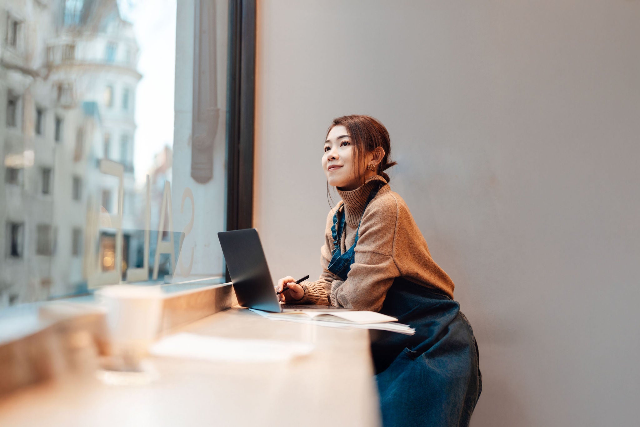 Young Asian woman looking through store window while working with laptop in cafe. Small business and young entrepreneur.