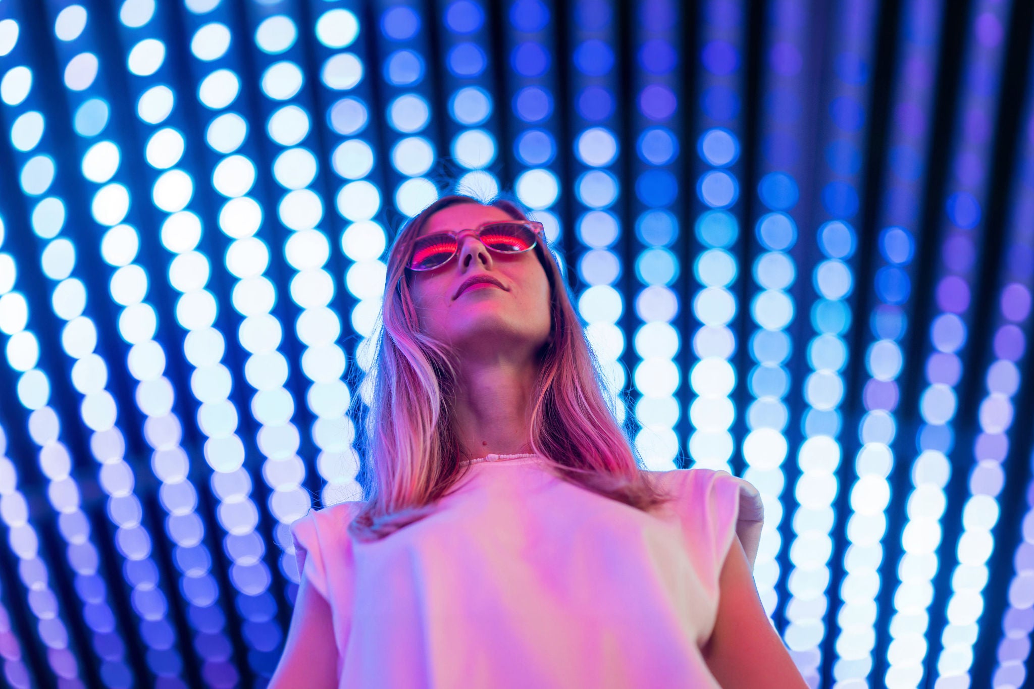 Teen hipster girl in stylish glasses standing on blue tunnel with neon light wall background, female teenager fashion model pretty young woman looking at night club city light glow. High quality photo