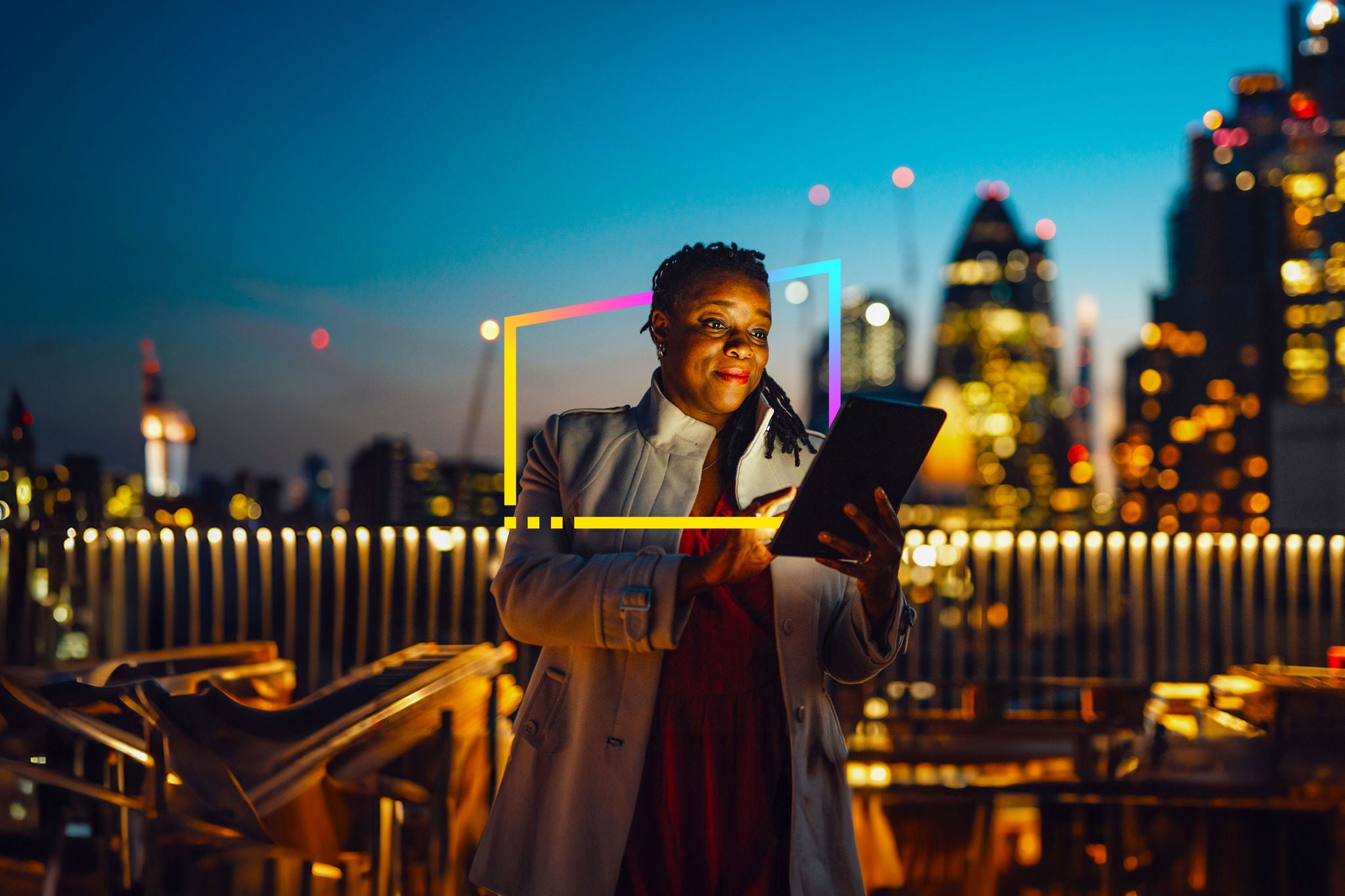 An adult African businesswoman using digital tablet at rooftop in business building against illuminated financial buildings and London cityscape at night