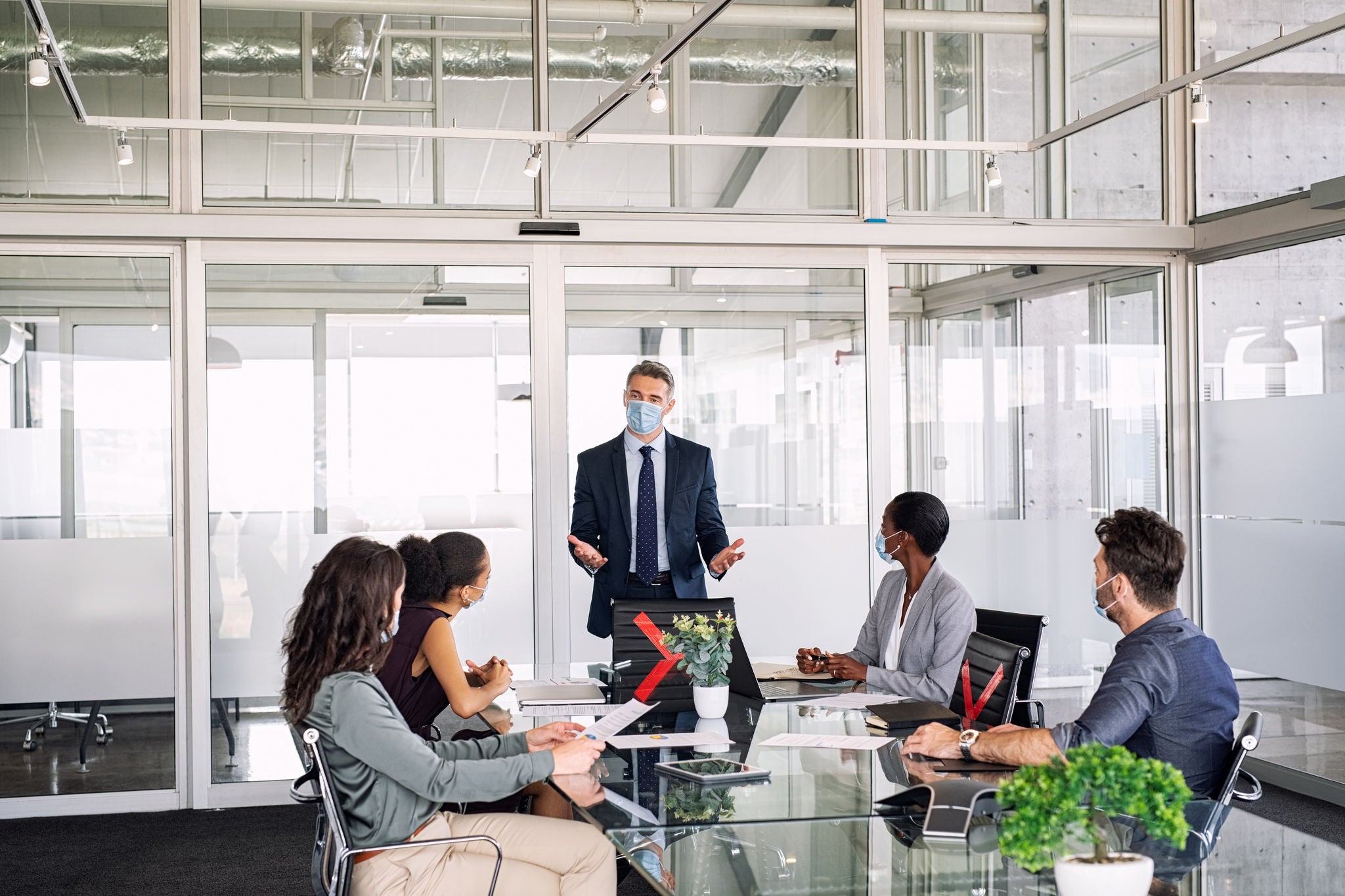 Mature manager in office discussing strategy with employees while wearing face mask during covid-19 pandemic. Group of multiethnic business people in meeting keeping social distancing in boardroom. Businessman and businesswomen working at modern corporate office keeping prevention against corona virus pandemic.