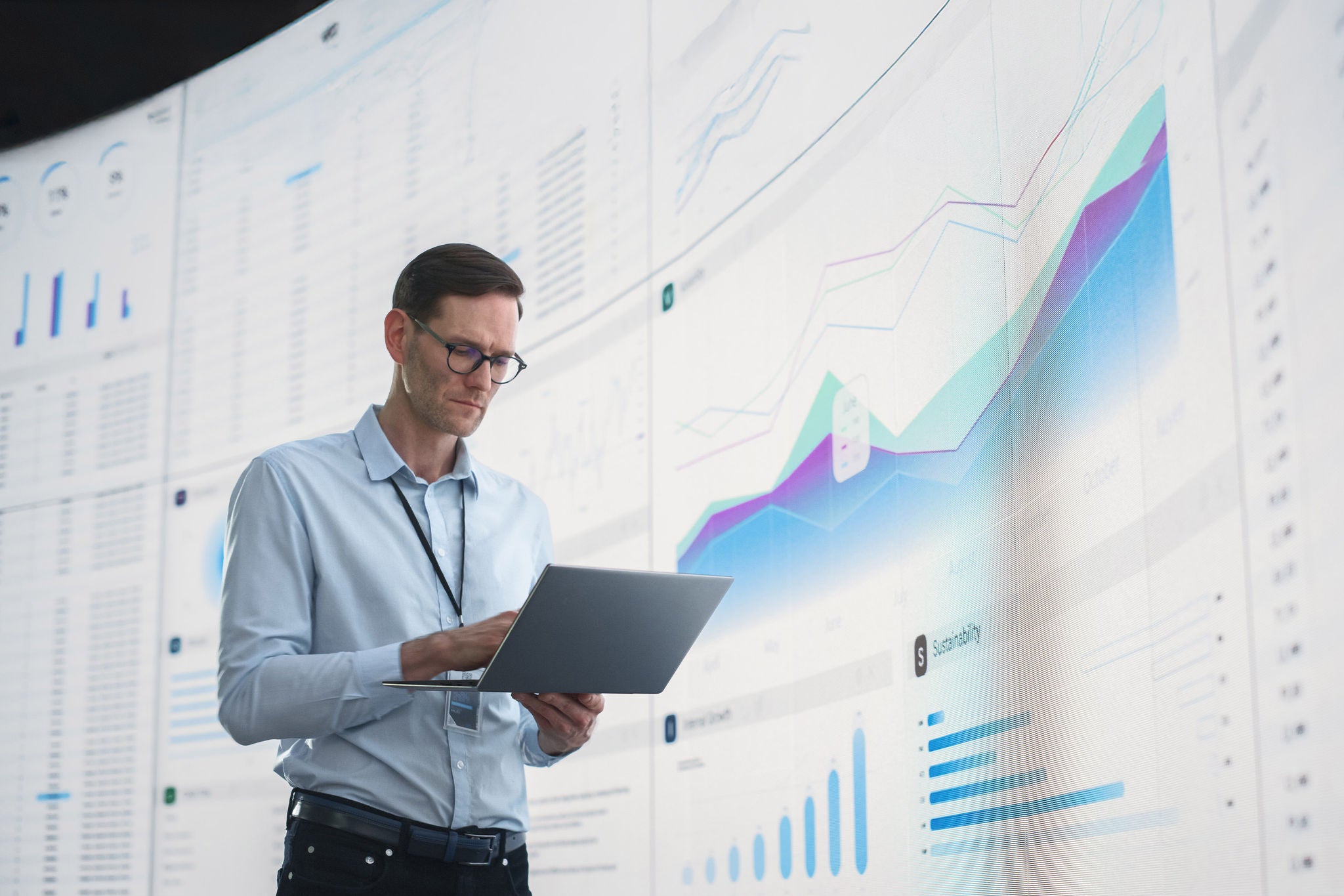 In The Office: Male Monitoring Specialist Holding Laptop Computer And Standing Next To Big Digital Screen With Graphs And Charts. Caucasian Man Analyzing Data, Performance Records, And Filing Report.