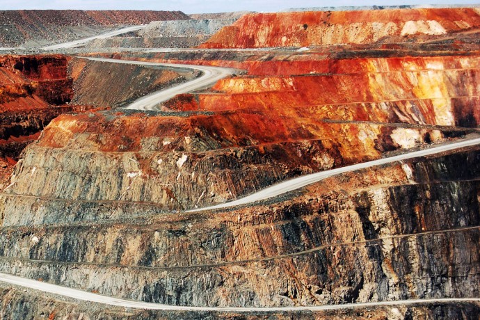 A vast open-pit mine featuring terraced layers of exposed earth in various shades of red, orange, brown, and gray