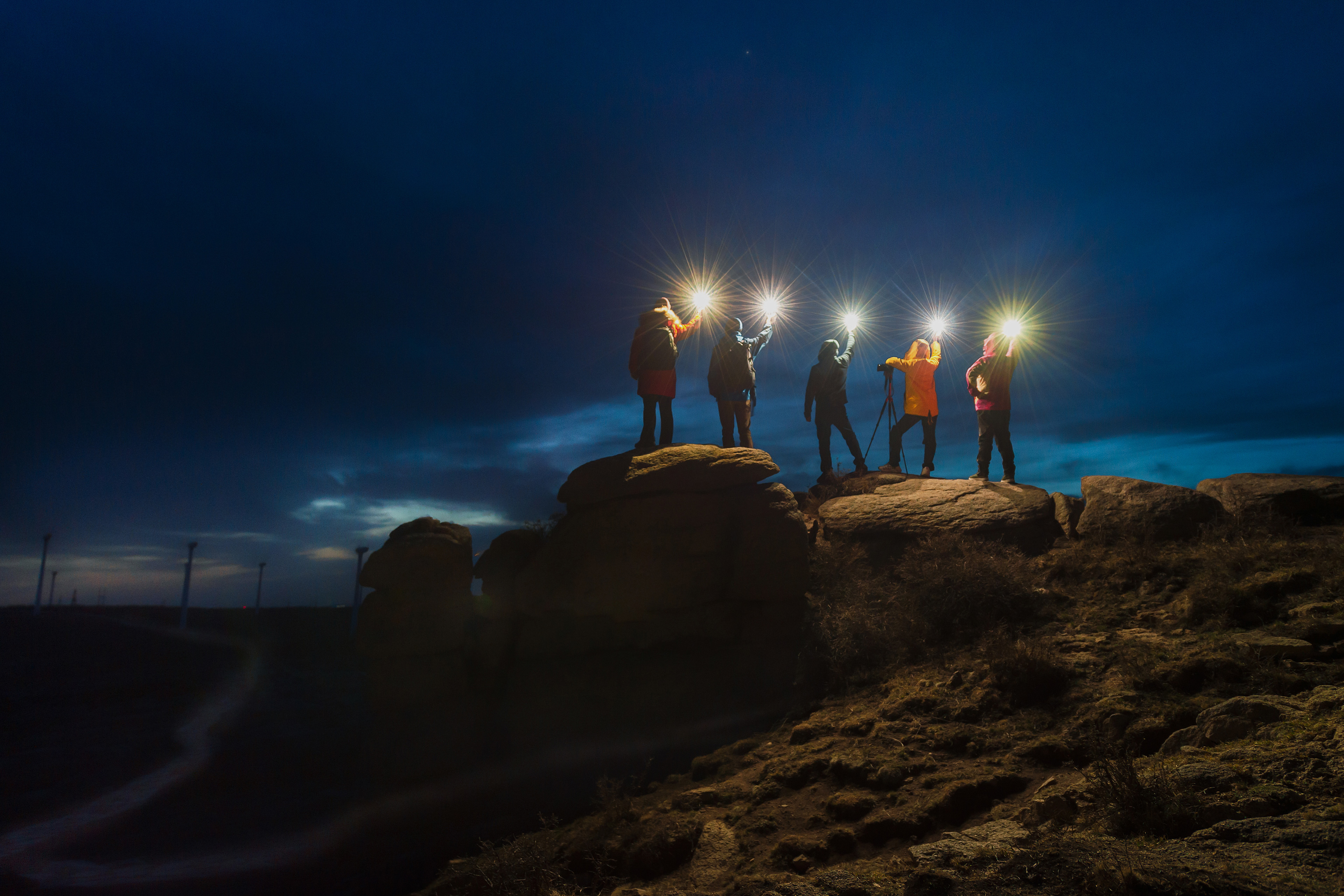 A group of people standing on a rocky hilltop at dusk, holding bright lights that illuminate the dark sky around them