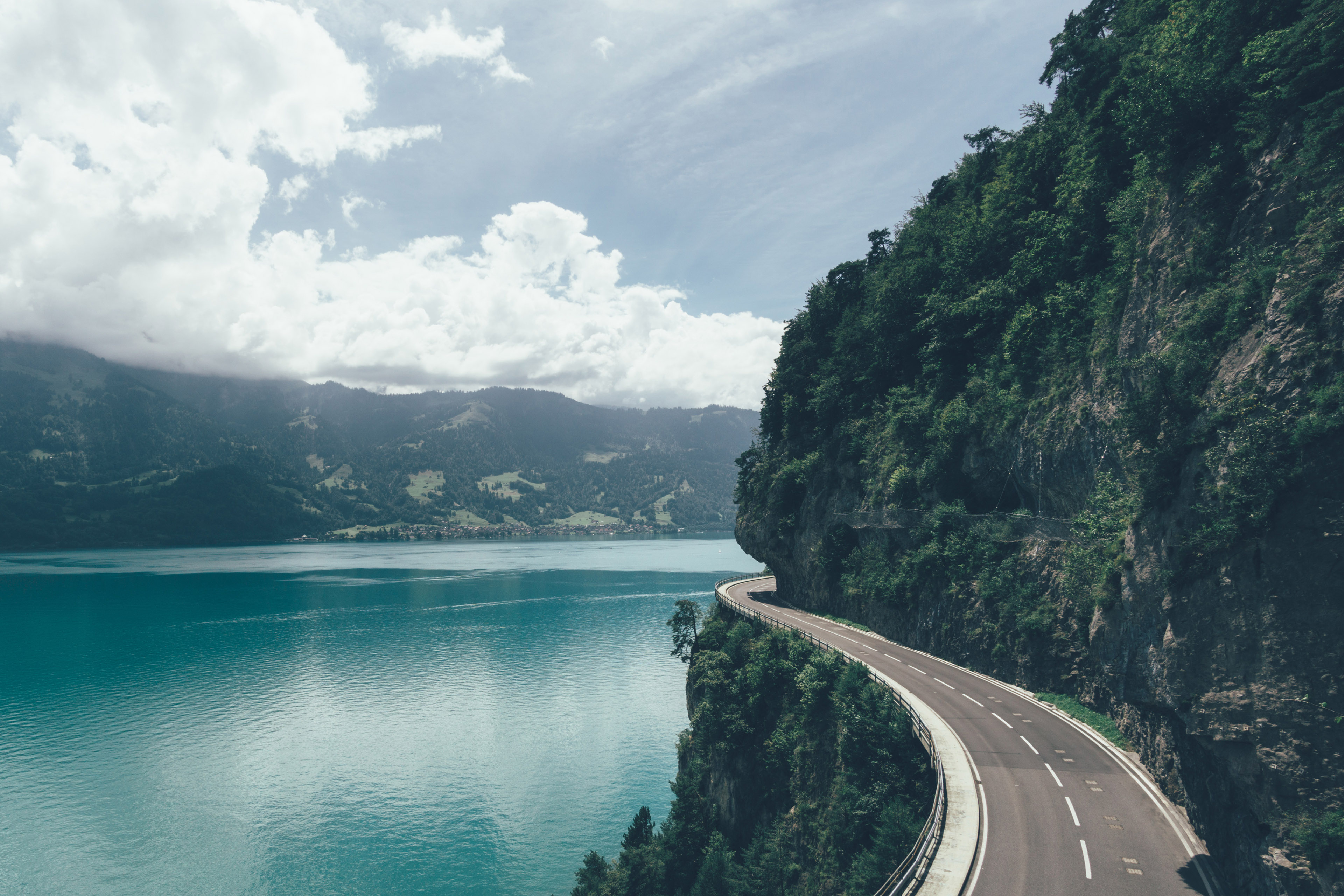 A scenic road curves along the edge of a cliff overlooking a calm, turquoise lake