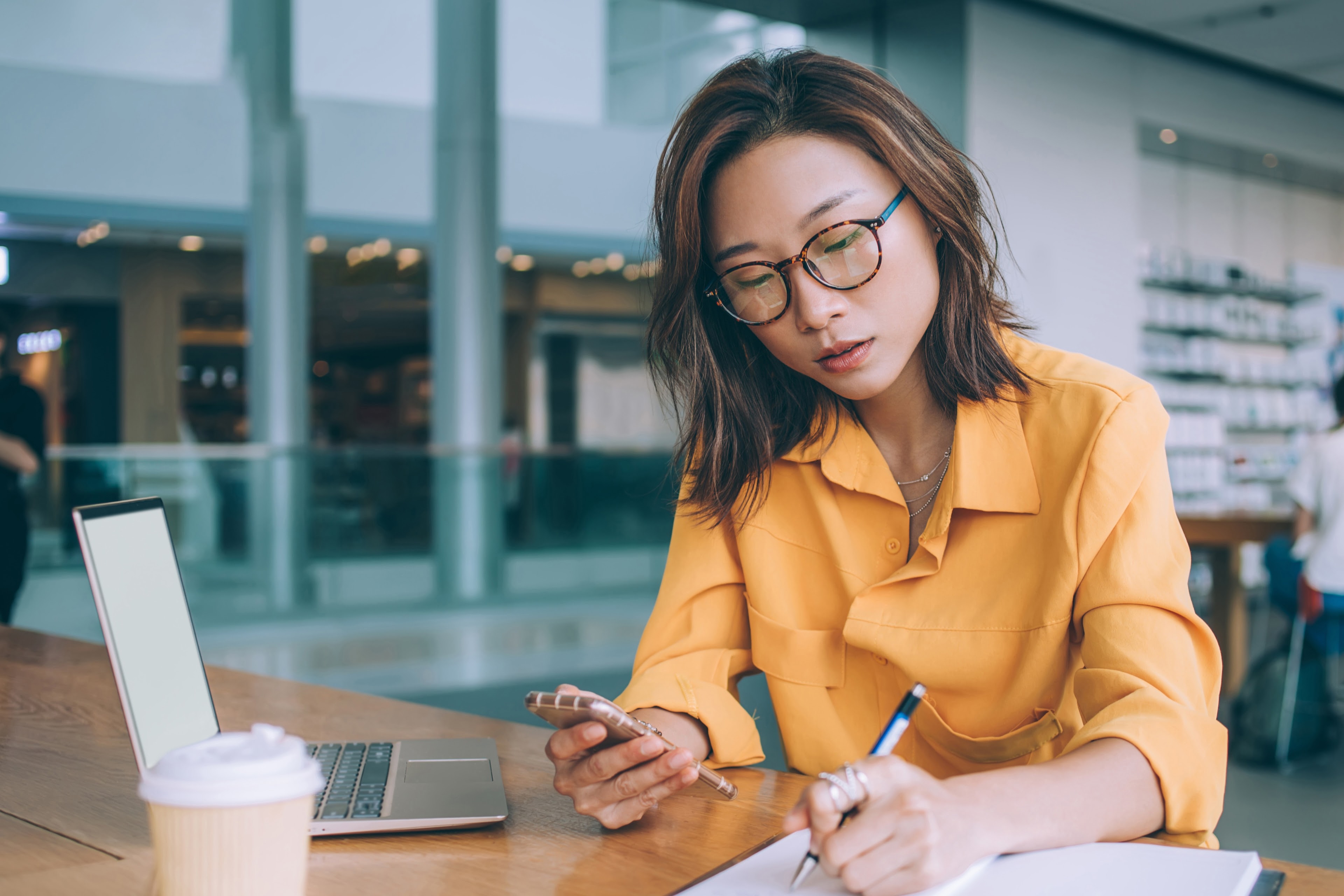 Thoughtful woman writing notes while browsing phone