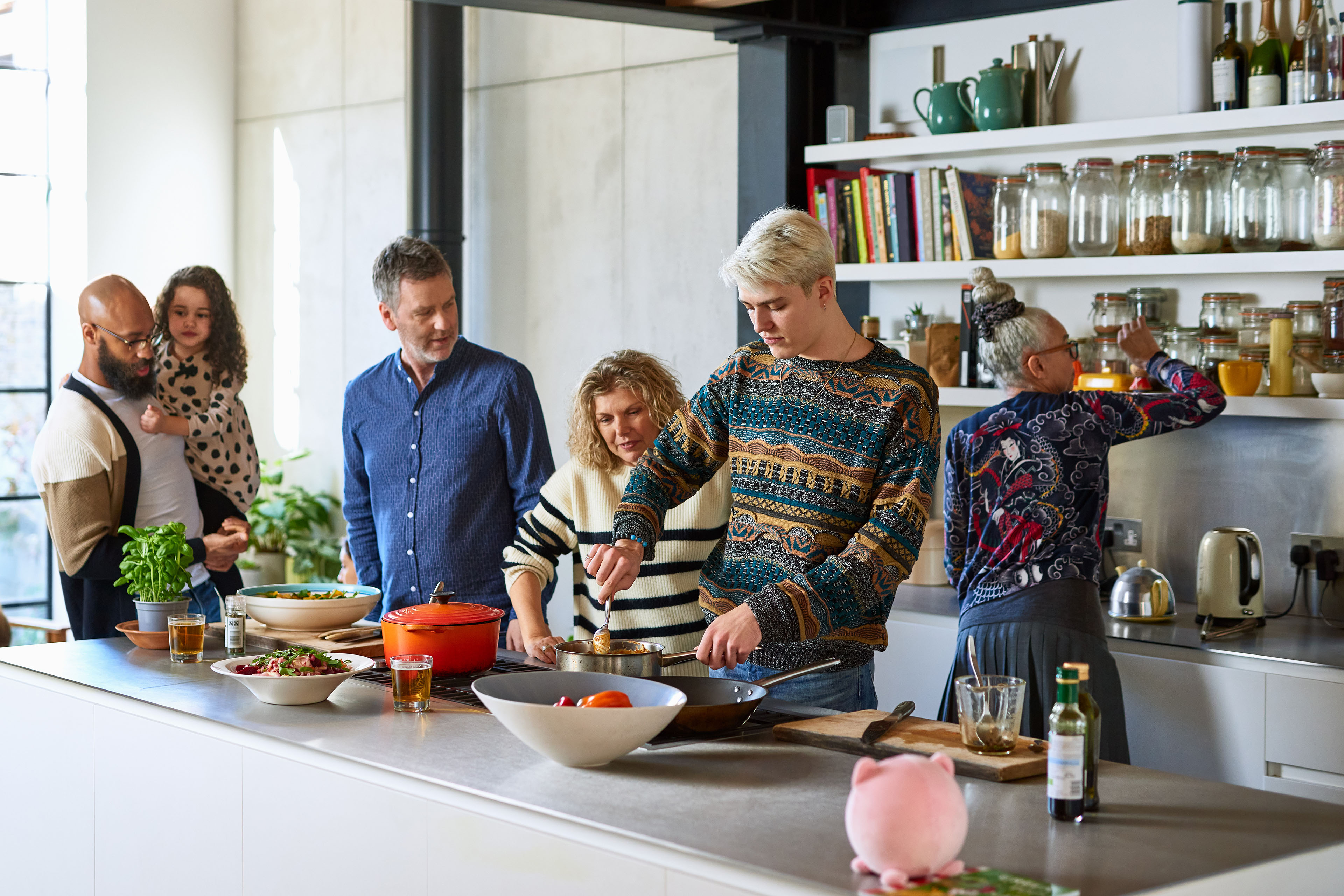 A photograph of a young man making a meal for an extended multiracial family