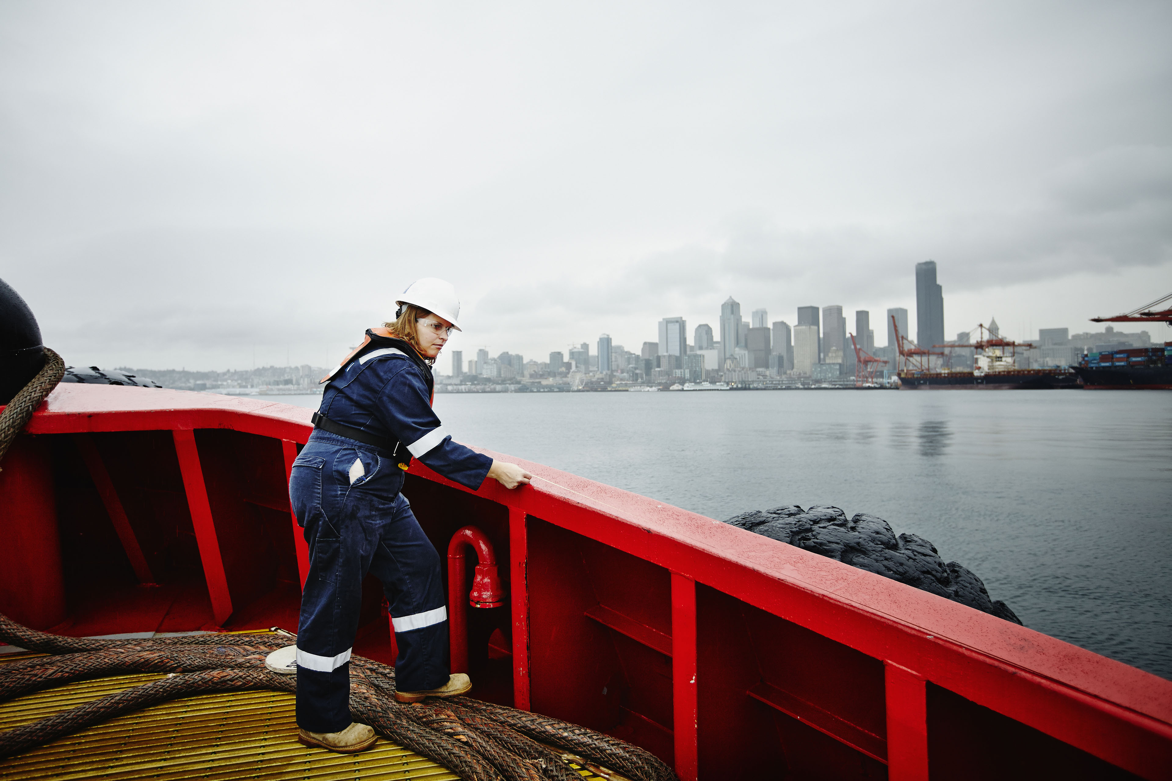 Female naval architect in hardhat and coveralls taking measurement along bulwark of commercial tugboat