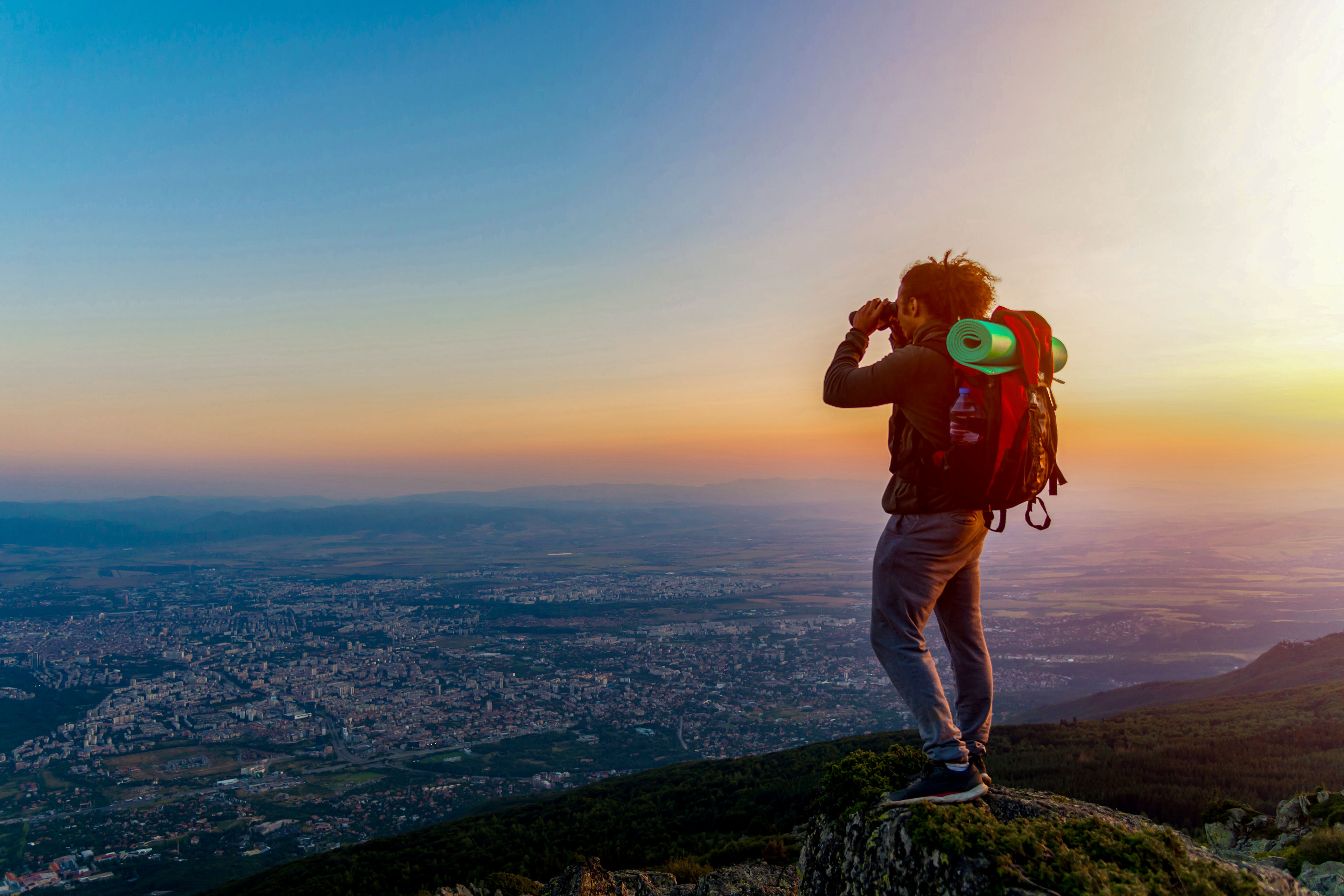 Man on a hilltop looking over a city with binoculars