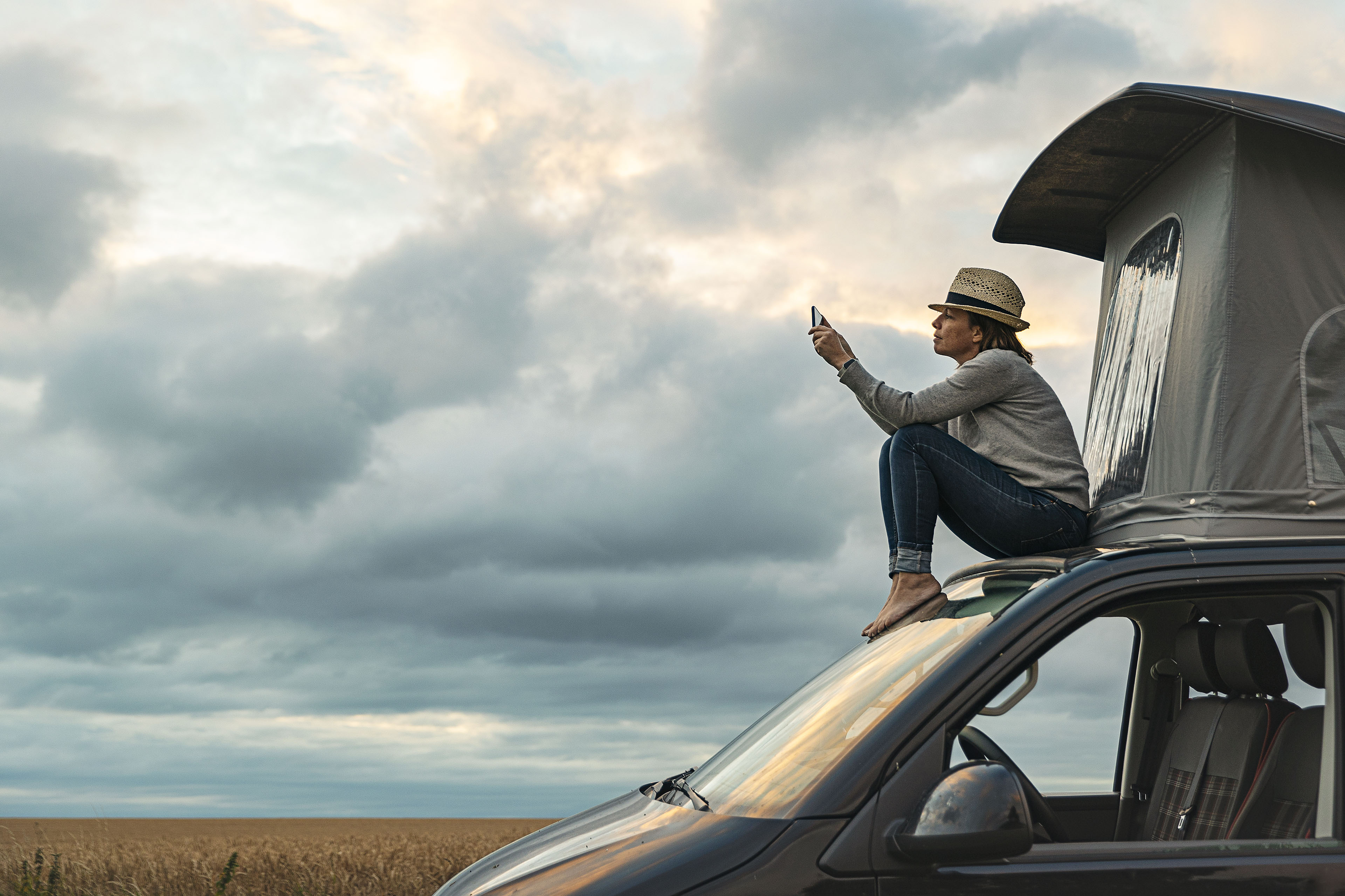 Woman taking a photo whilst sitting on roof of camper at sunset