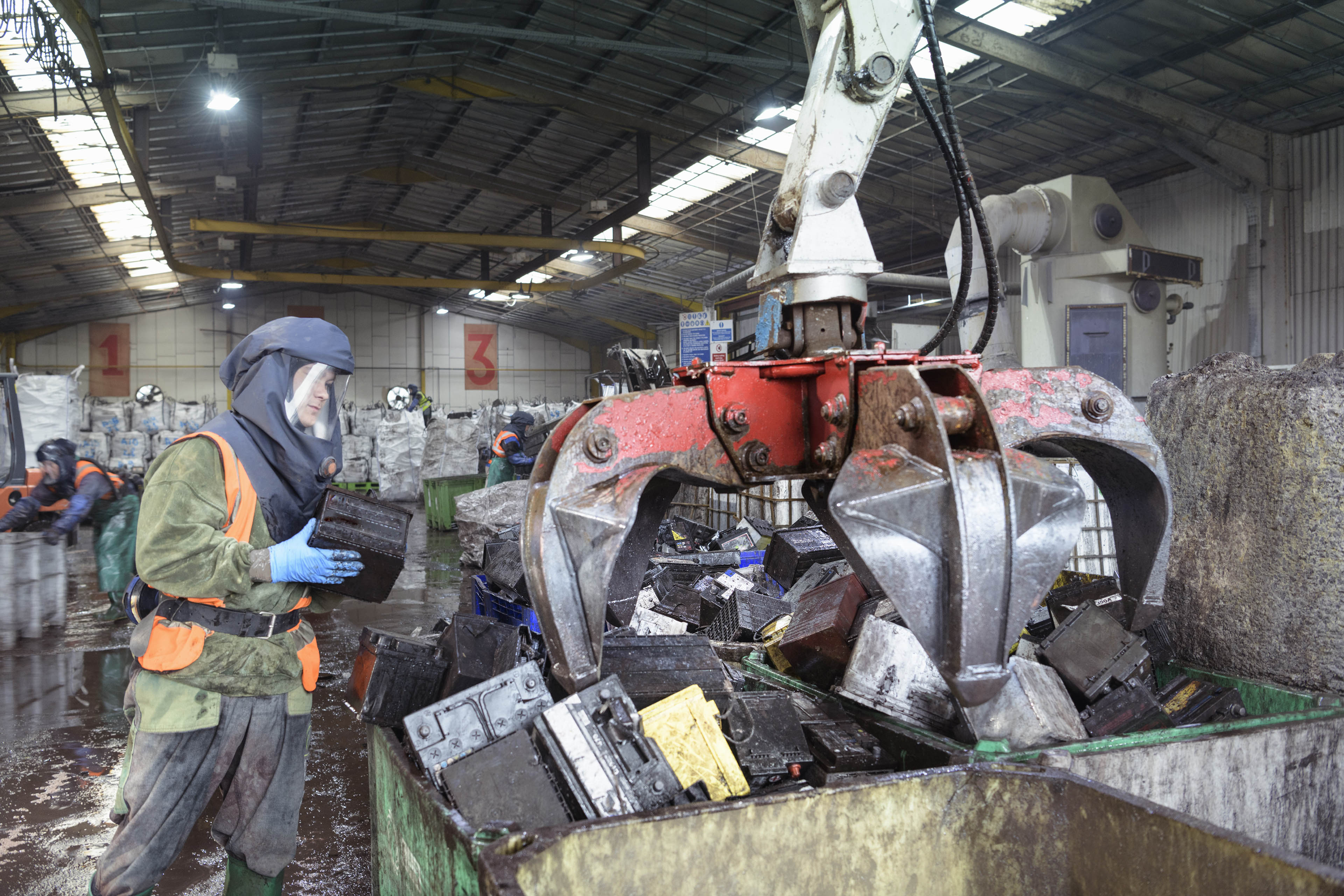 Worker in protective clothing loading vehicle batteries into grab bin in vehicle battery recycling plant